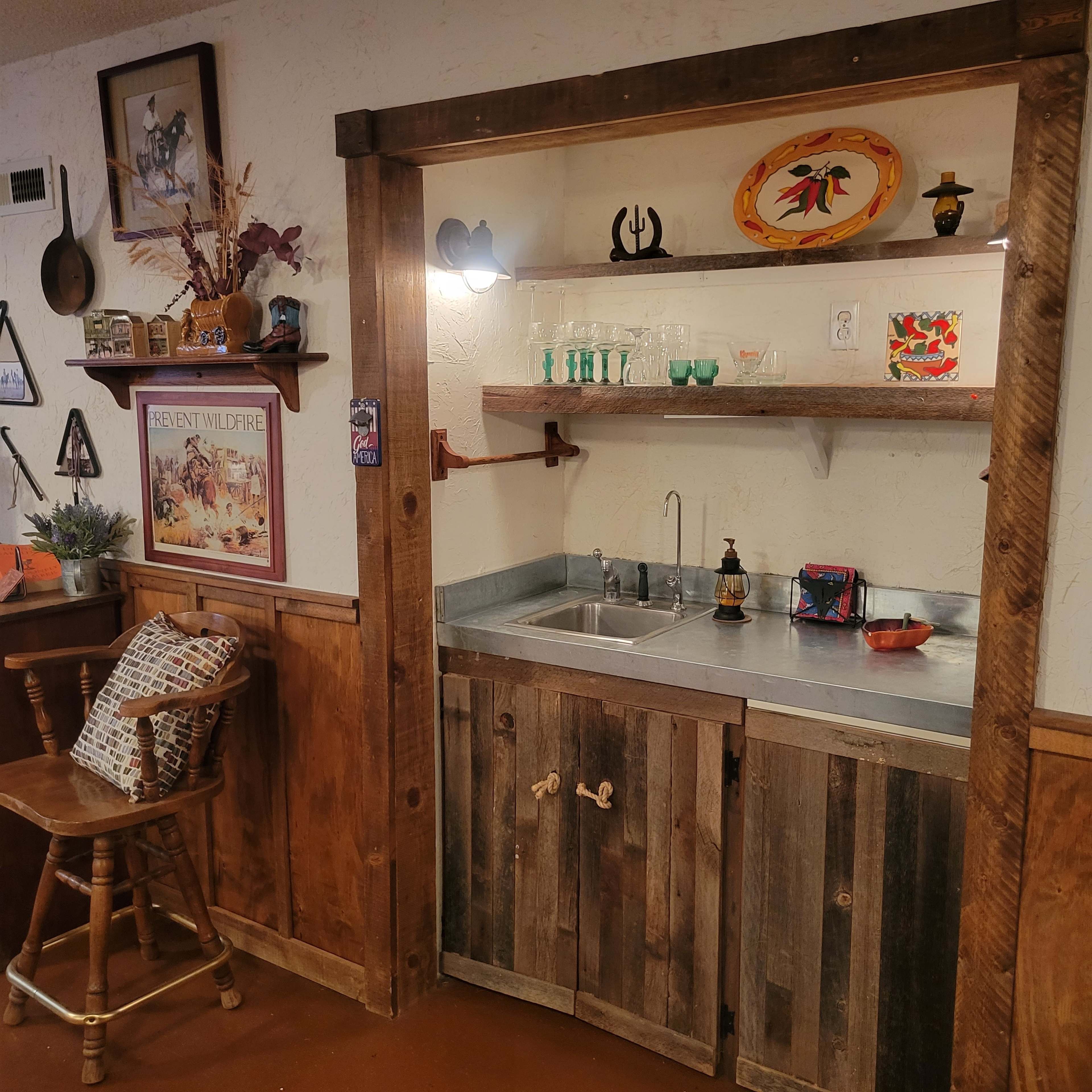 The image shows a rustic kitchenette with a metal sink, wooden cabinets, and decorative items displayed on the shelves.