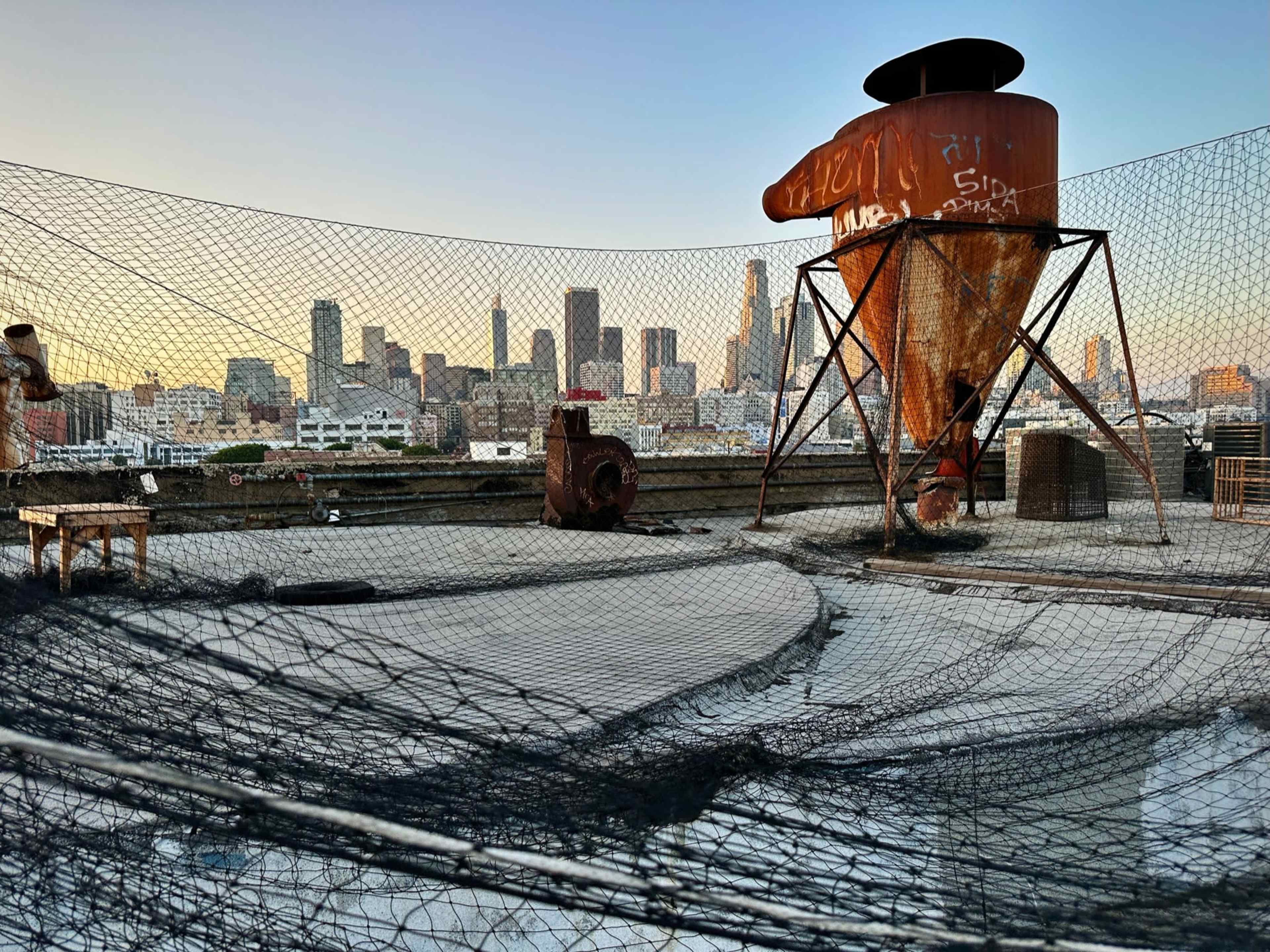 A rooftop view of a city skyline, featuring a rusted water tower and a netting structure in the foreground.