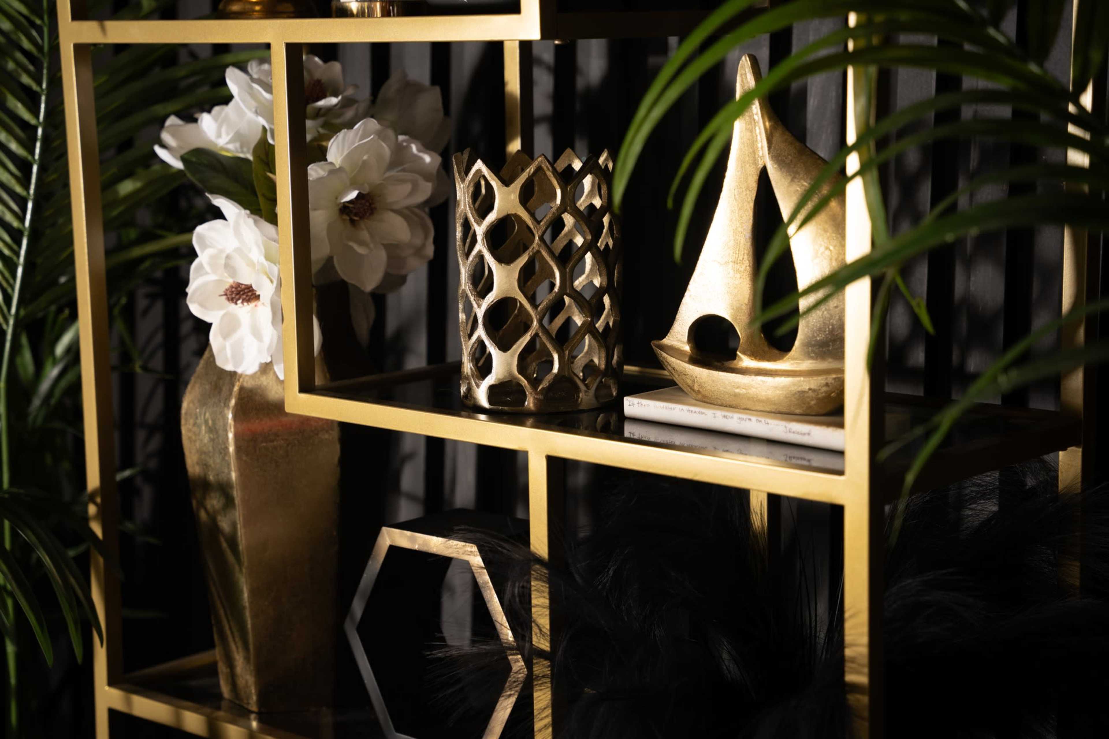 The image features a decorative shelf with gold accents, showcasing a vase with white flowers, a patterned candle holder, a decorative sailboat, and a geometric object, all framed by green leaves.
