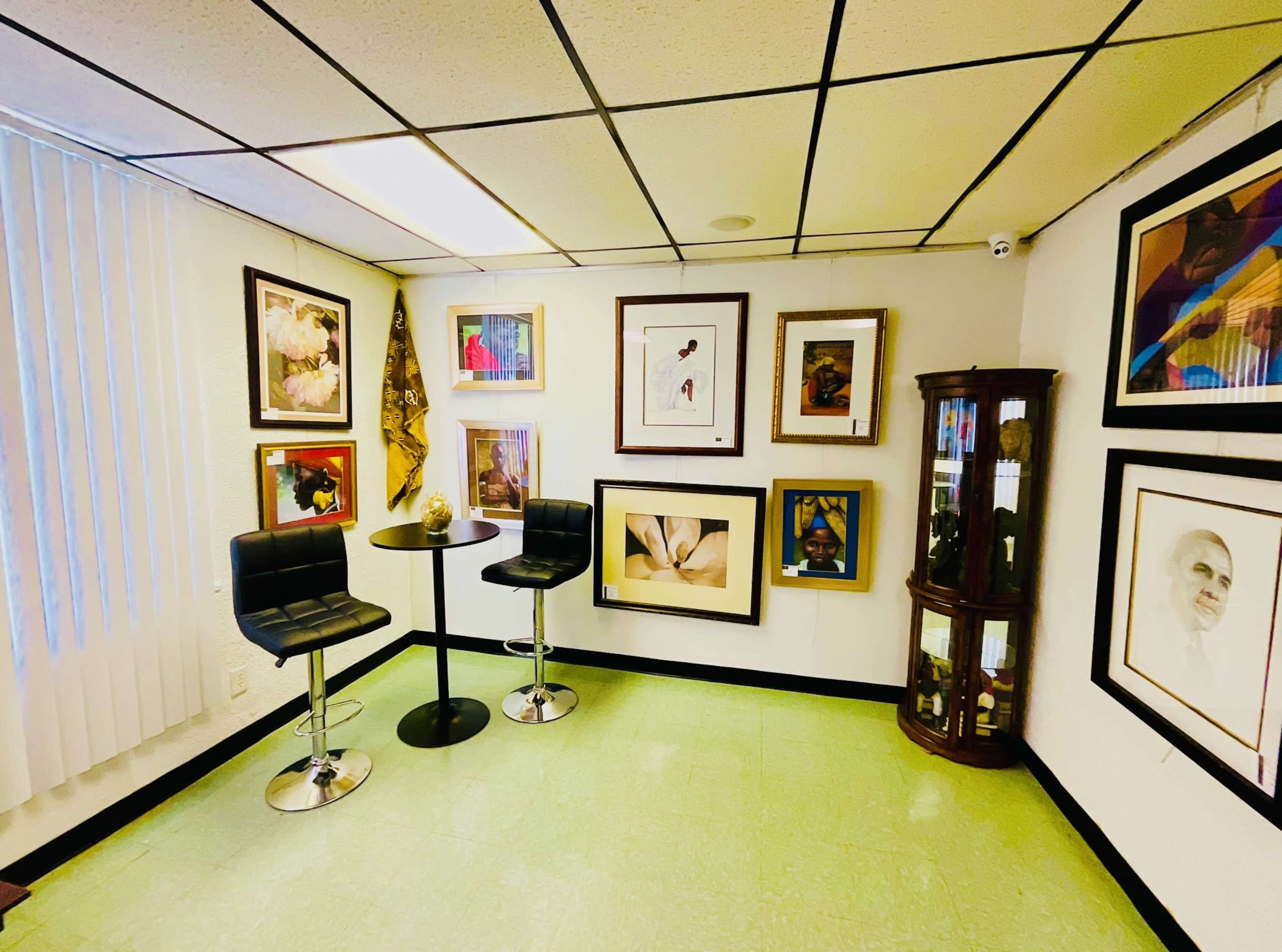 A small gallery room features a set of framed artworks on the walls, a round table with two black stools, and a display cabinet in one corner.
