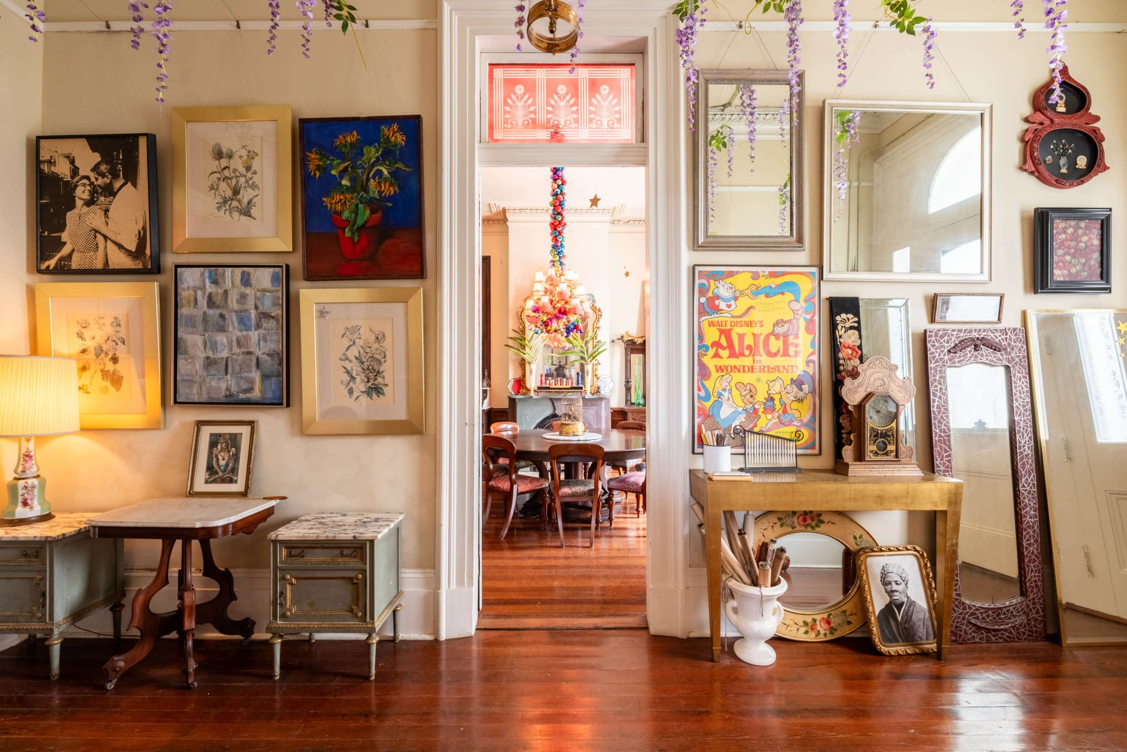 A hallway adorned with various framed artworks, a lamp, and mirrors, leading to a dining area with a colorful chandelier.