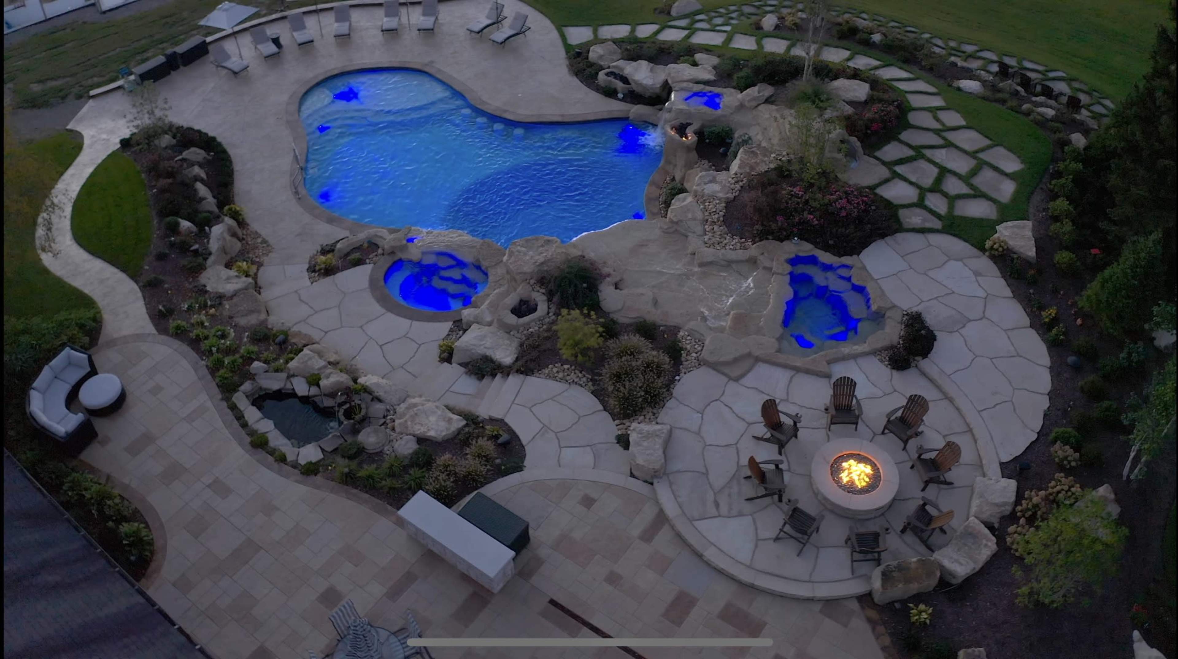 An aerial view shows a landscaped pool area with a blue swimming pool, stone pathways, and a fire pit surrounded by chairs.