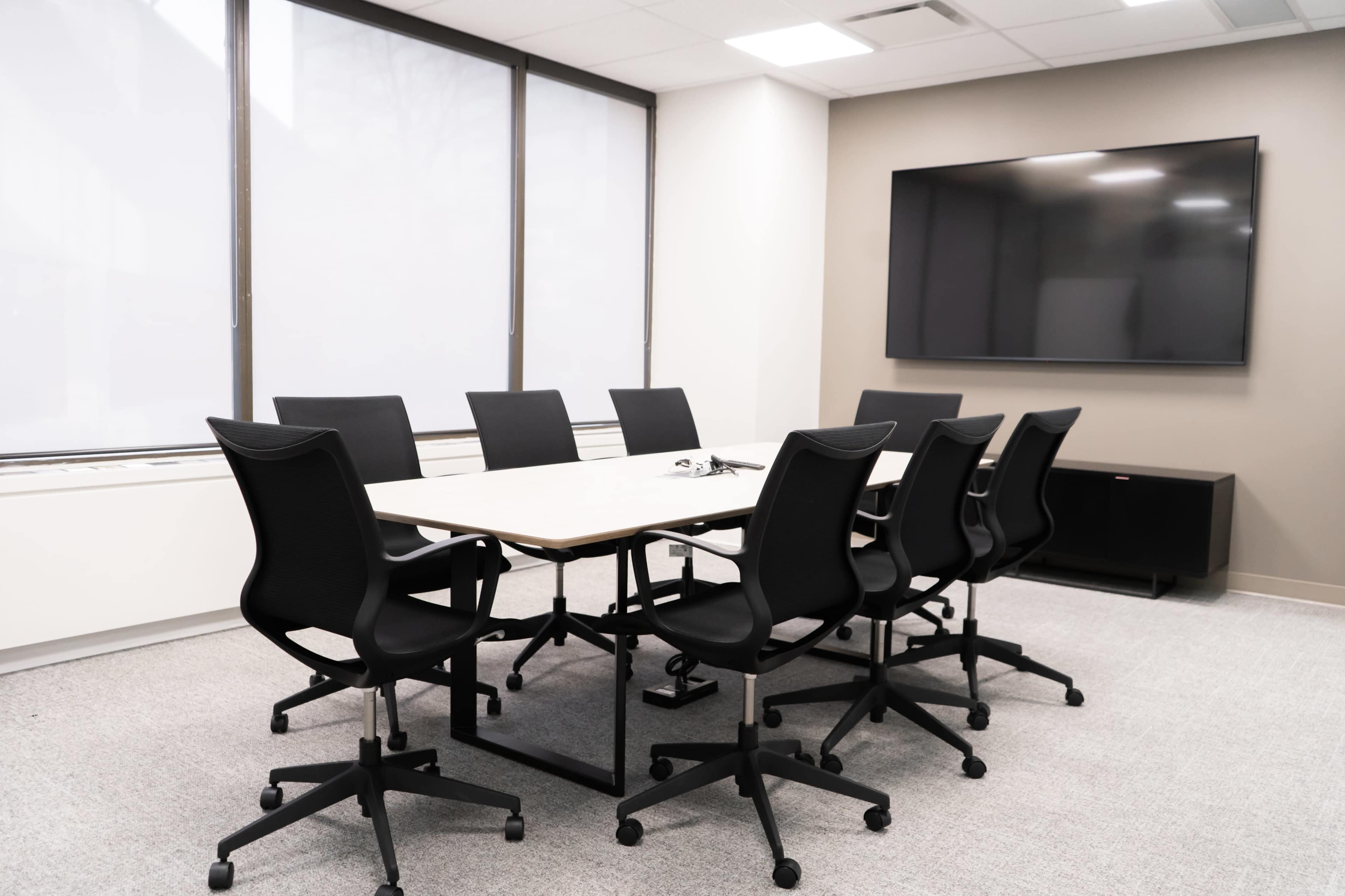 A meeting room features a long table surrounded by eight black swivel chairs, with a large television mounted on the wall.