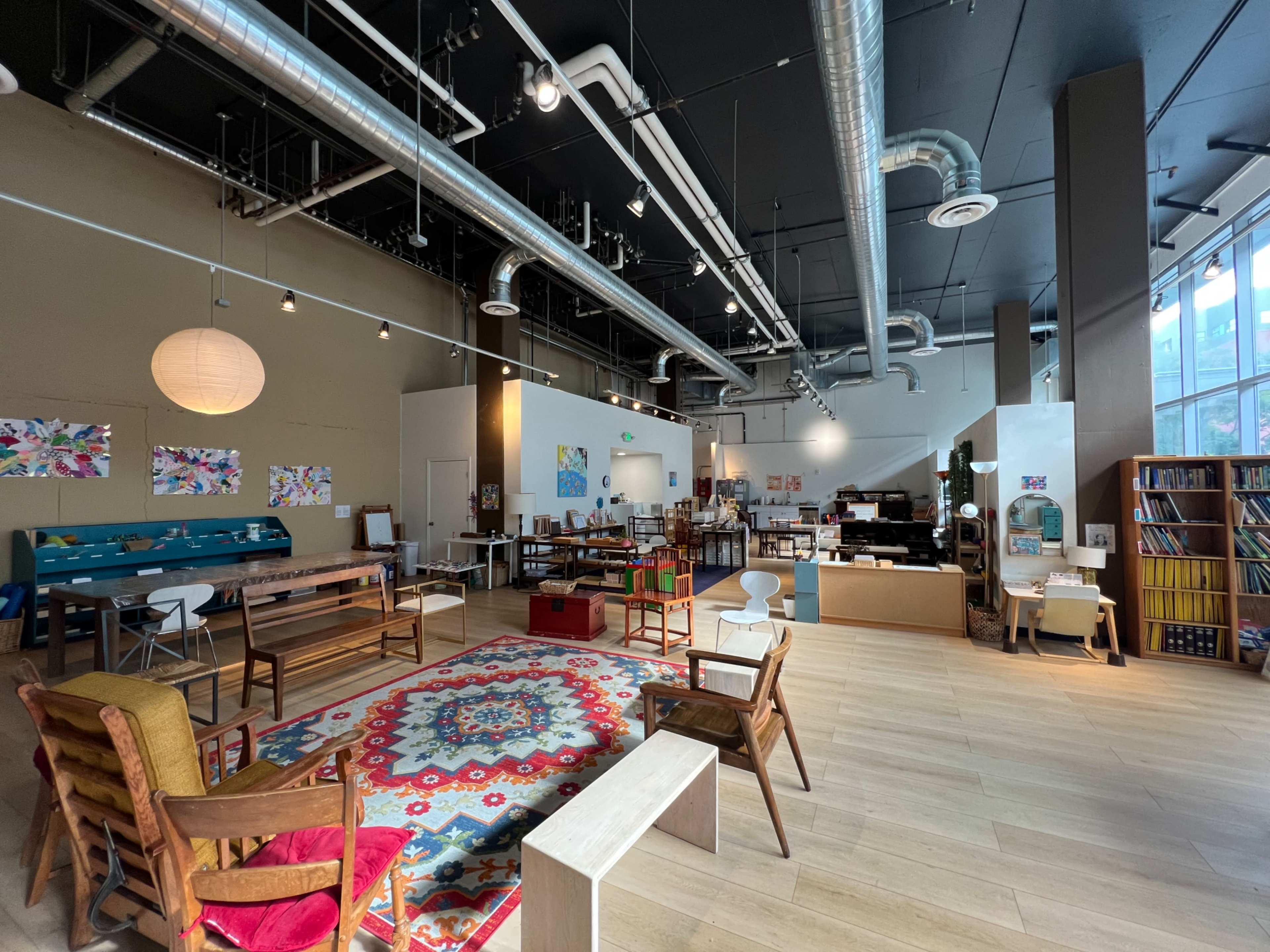 The image shows a spacious, well-lit room with various tables, chairs, and shelves arranged in a modern design, featuring colorful artwork on the walls.