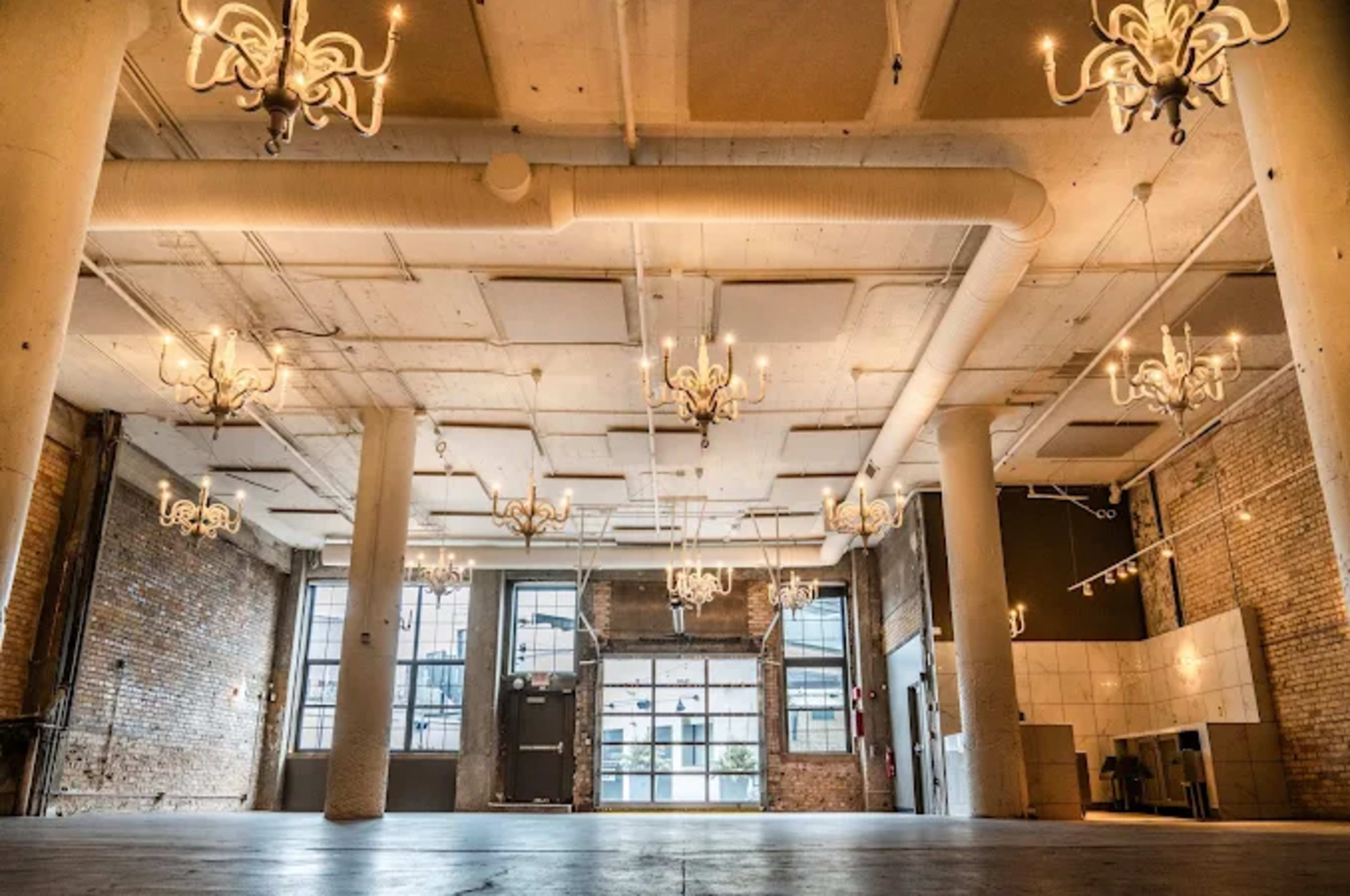 The image shows a spacious room with brick walls, large windows, and multiple chandeliers hanging from the ceiling.