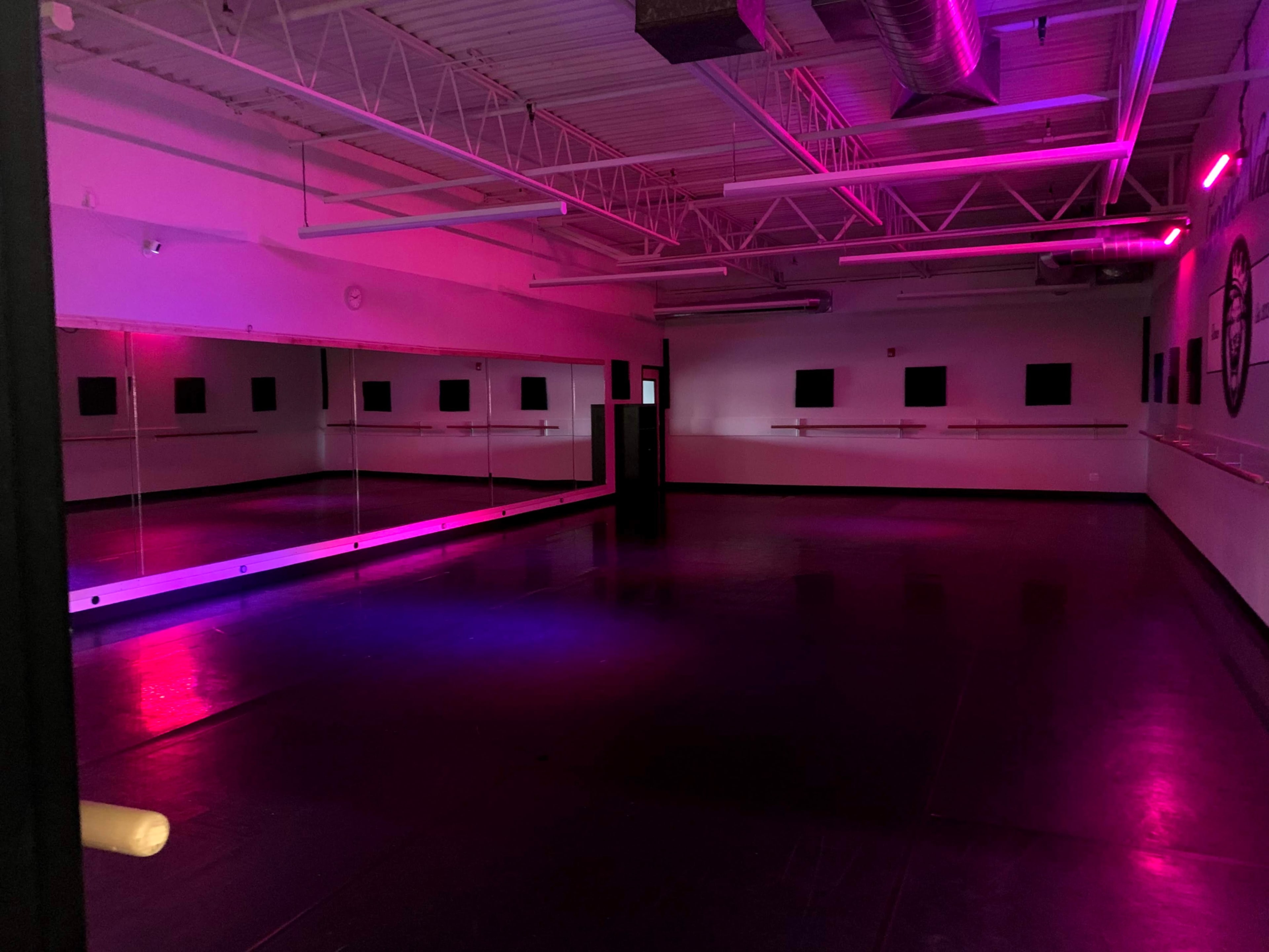An empty dance studio features a mirrored wall and soft pink and purple lighting.