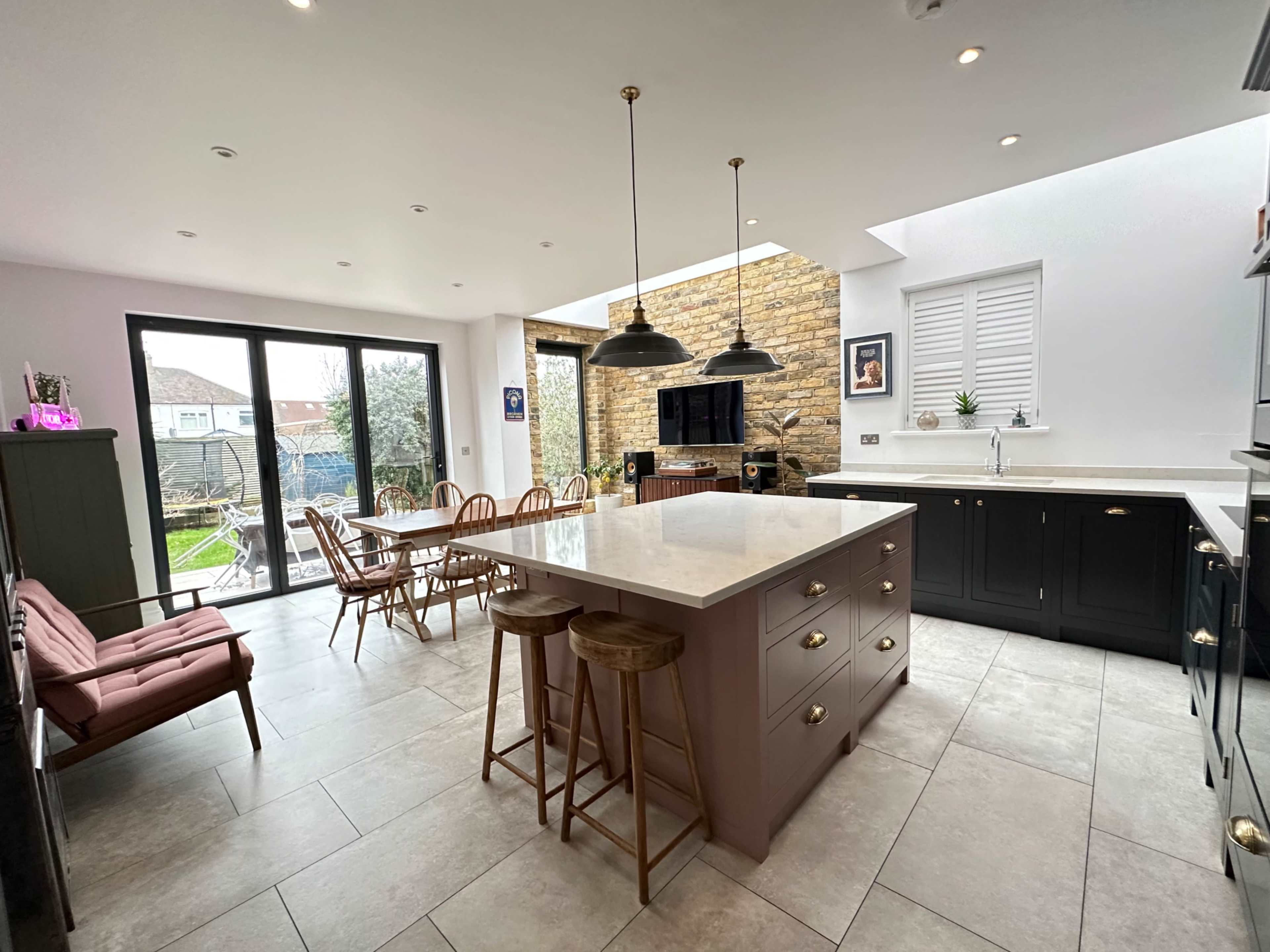 The image shows a modern kitchen featuring an island with bar stools, a dining area with wooden chairs, and large glass doors leading to a garden space.