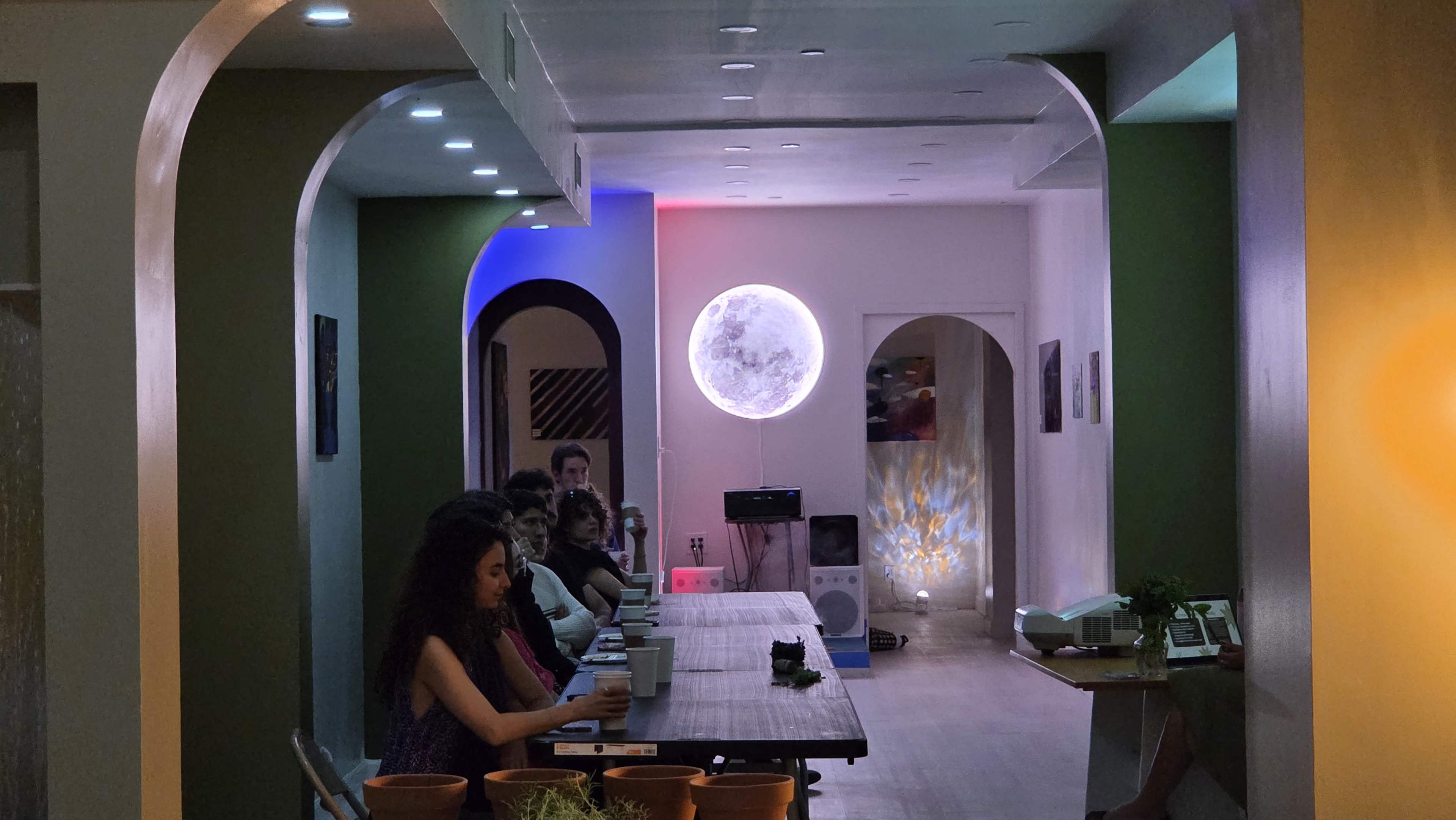 The image shows a modern indoor space with a long table and several people seated, illuminated by colorful lighting and featuring a large moon-shaped light on the wall.