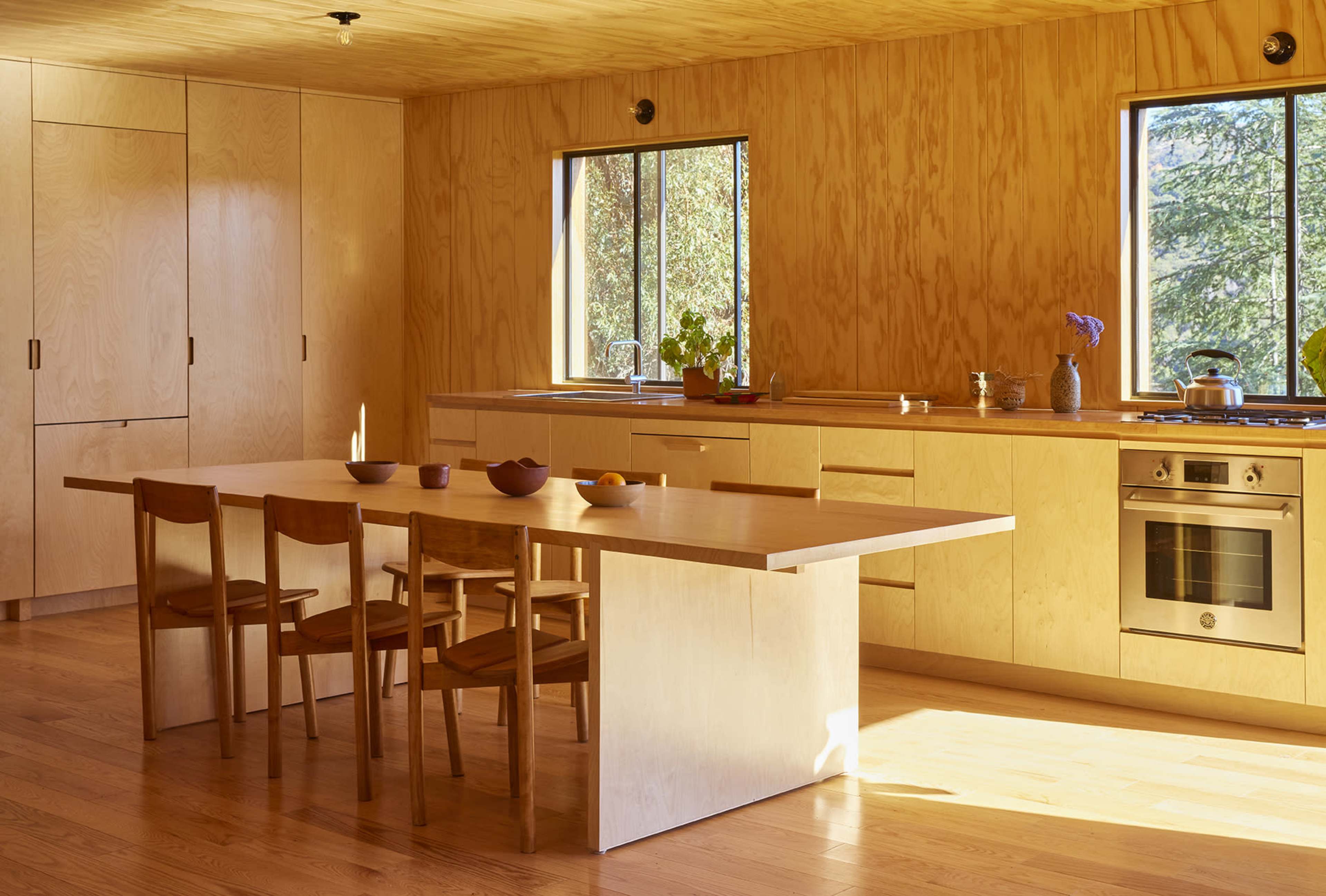 A modern kitchen with wooden cabinetry, a large table, and four chairs, illuminated by natural light from two windows.
