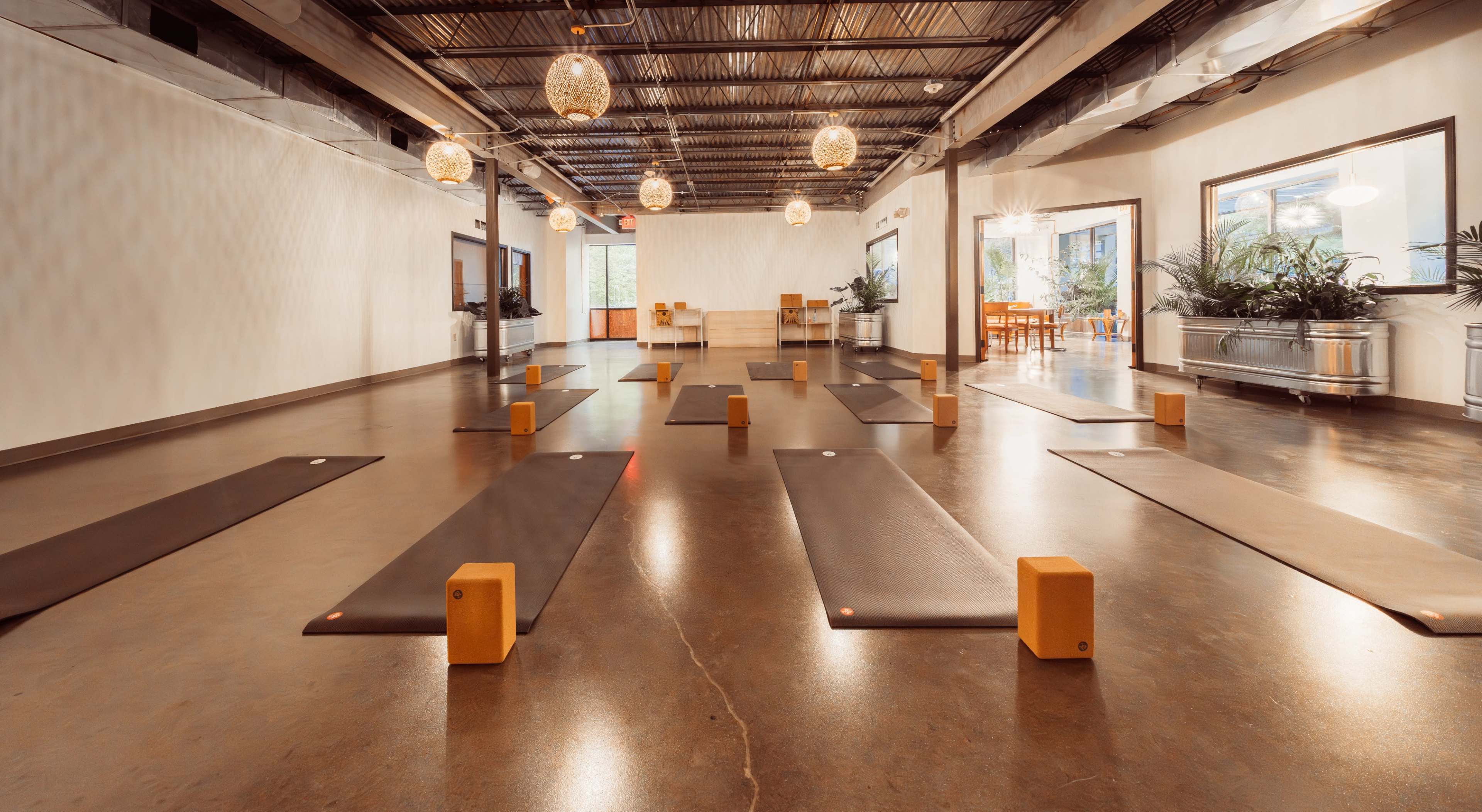 The image shows a spacious yoga studio with multiple yoga mats arranged in rows, wooden blocks placed at the front of each mat, and decorative pendant lights hanging from the ceiling.