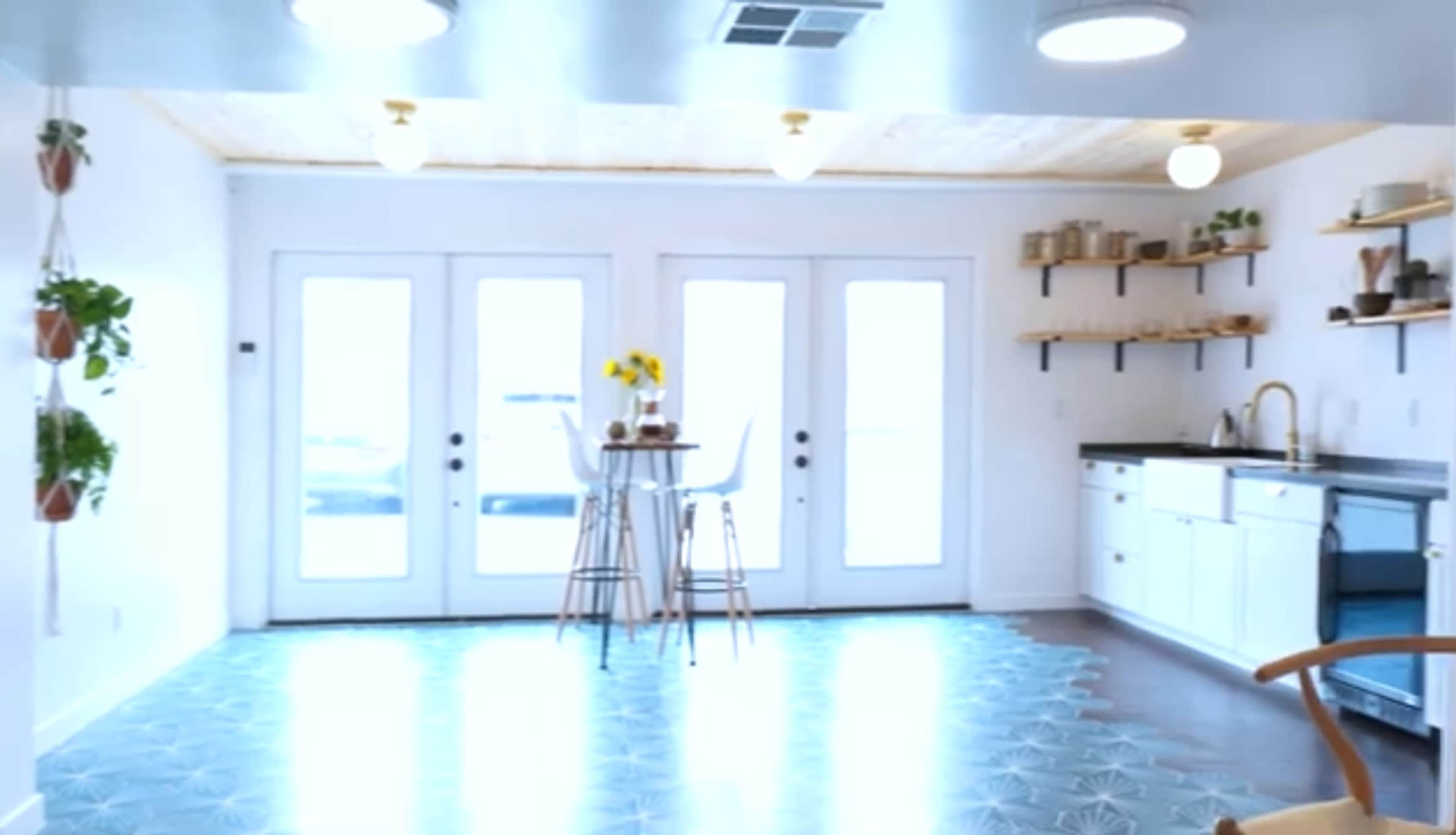 A bright, open kitchen and dining area features a small table with stools, plants hanging from the ceiling, and a modern kitchen layout with white cabinetry and shelves.