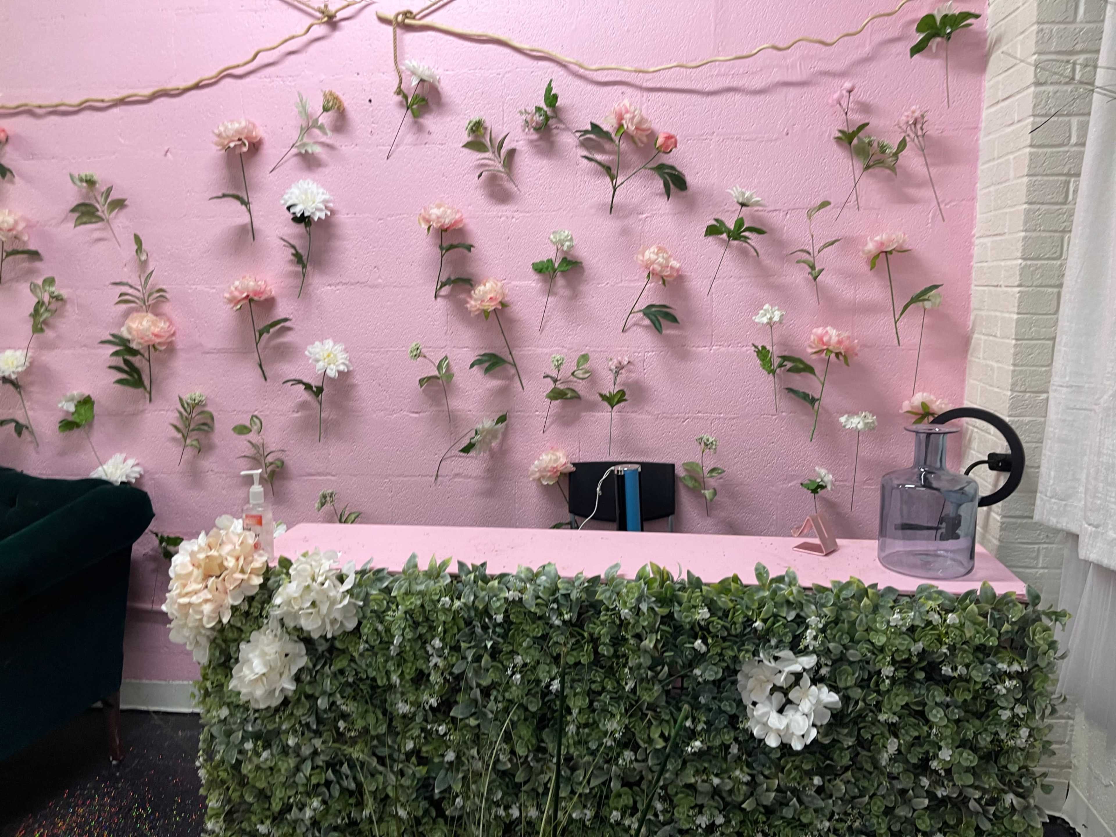 The scene features a pink wall adorned with fabric flowers and a reception desk covered in greenery, with a glass pitcher and decorative items on top.