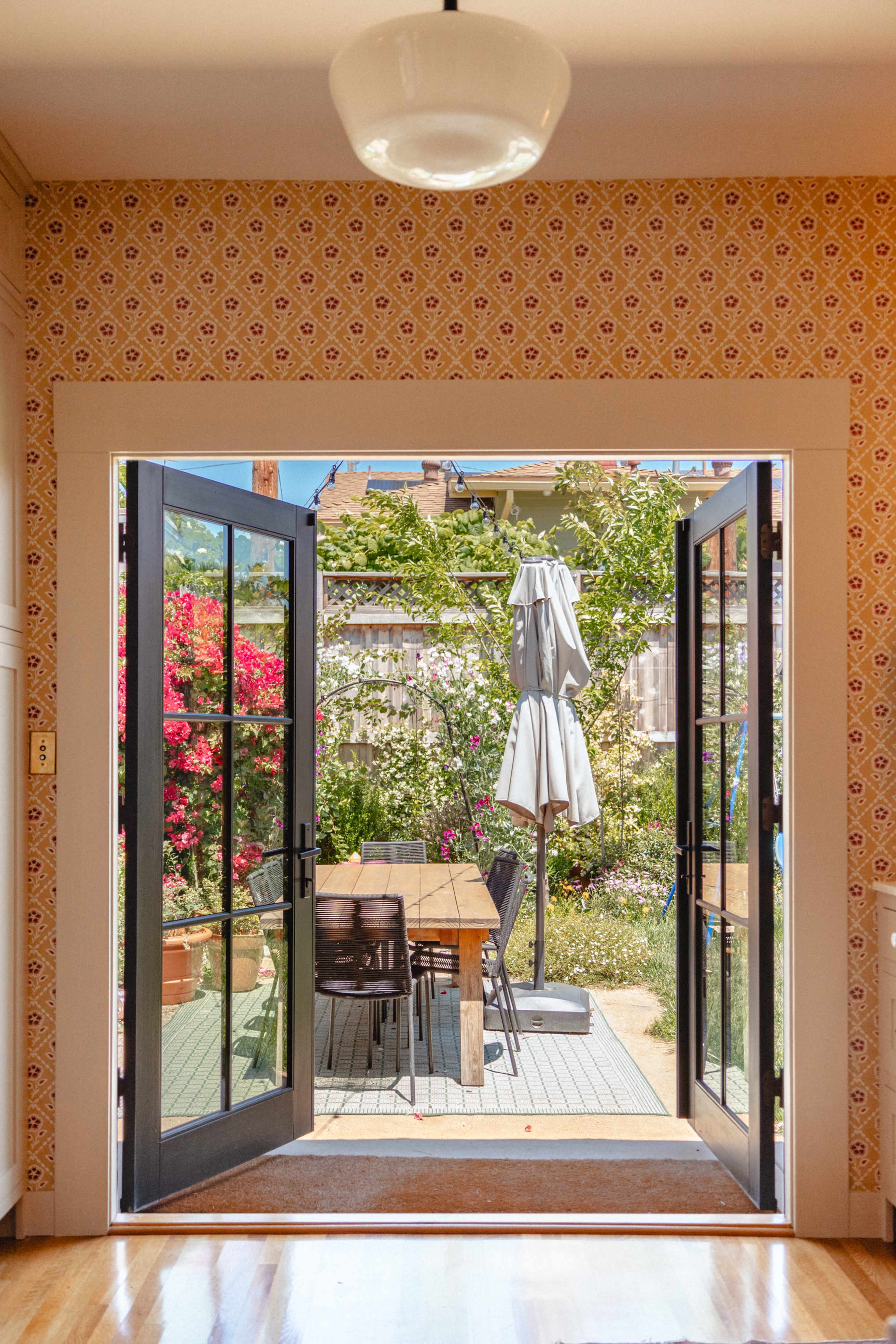 The image shows a spacious kitchen with double doors open to a patio featuring a wooden table and an umbrella surrounded by vibrant flowers and greenery.