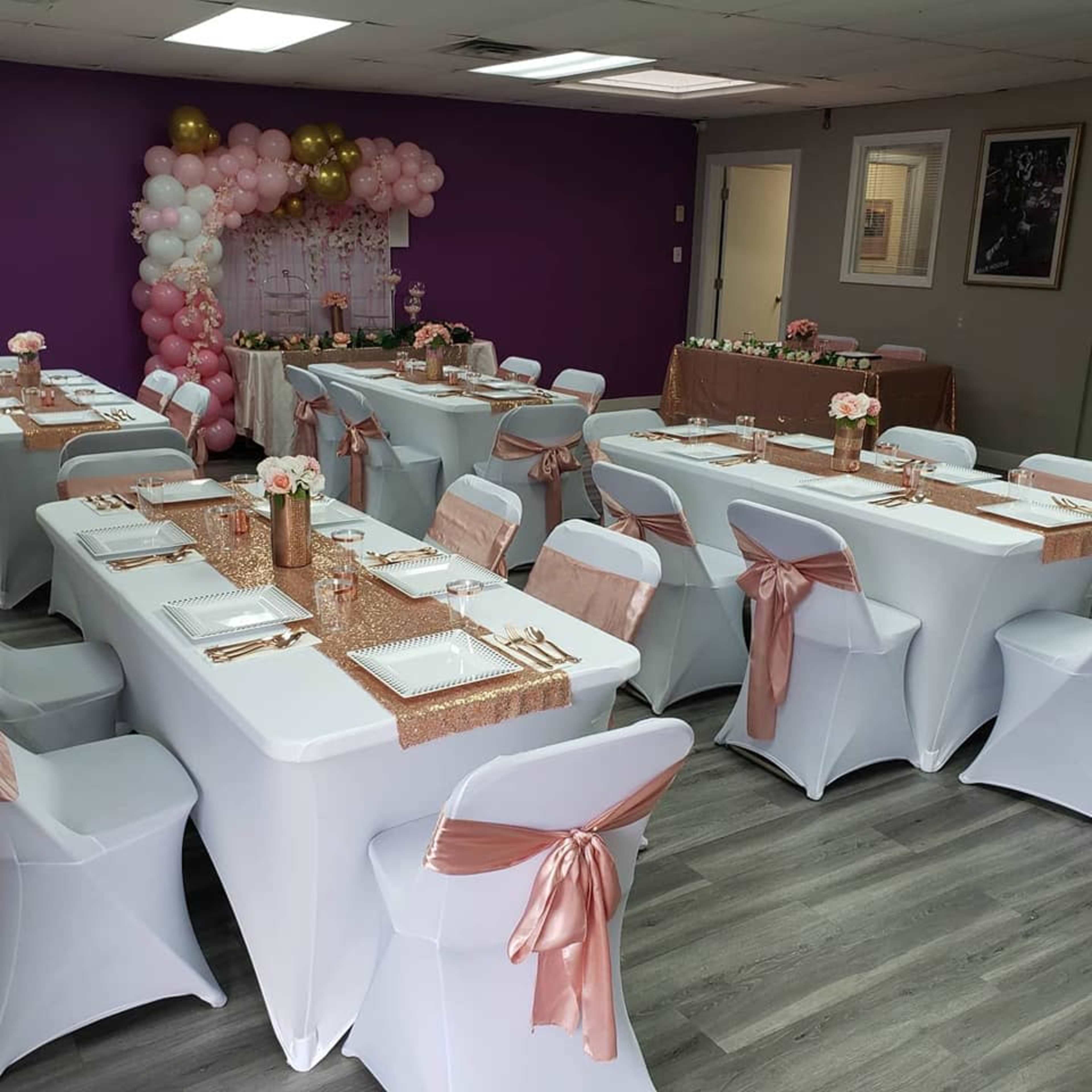 The image shows a decorated event space with several tables set with white tablecloths, each adorned with pink and gold accents, and a backdrop of balloons.