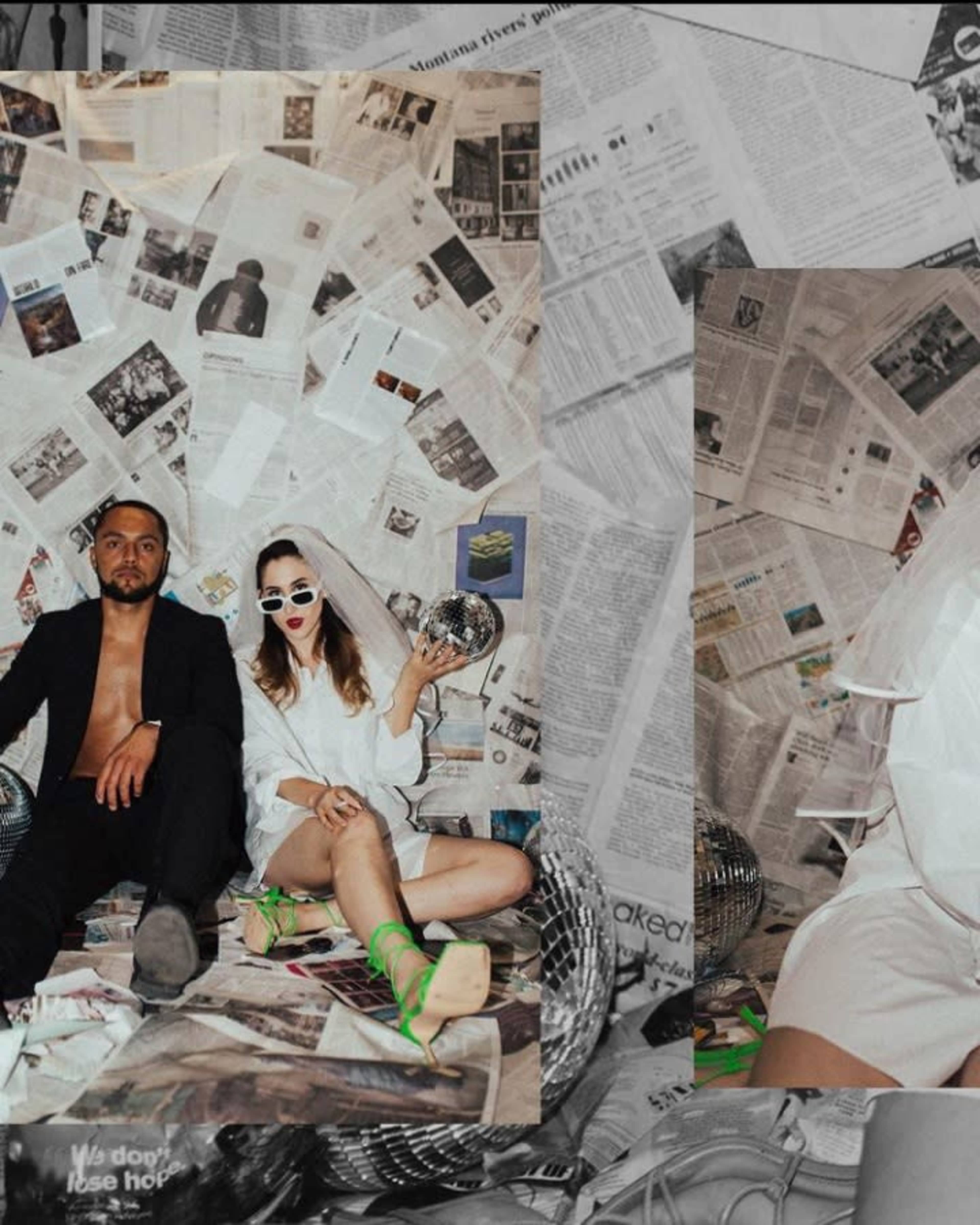 A man and a woman sit against a backdrop of newspapers, with the woman wearing sunglasses and holding a disco ball.