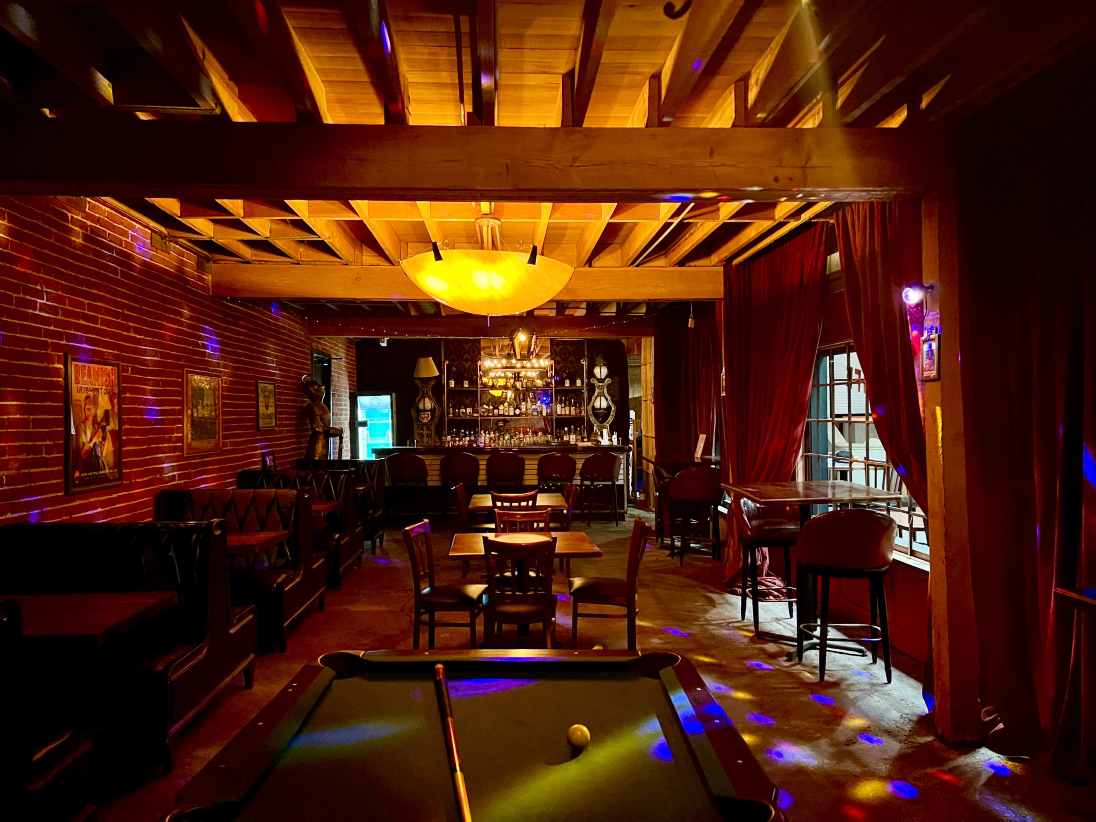 A dimly lit bar with a pool table in the foreground, surrounded by wooden furnishings and brick walls, and soft lighting from a ceiling fixture.
