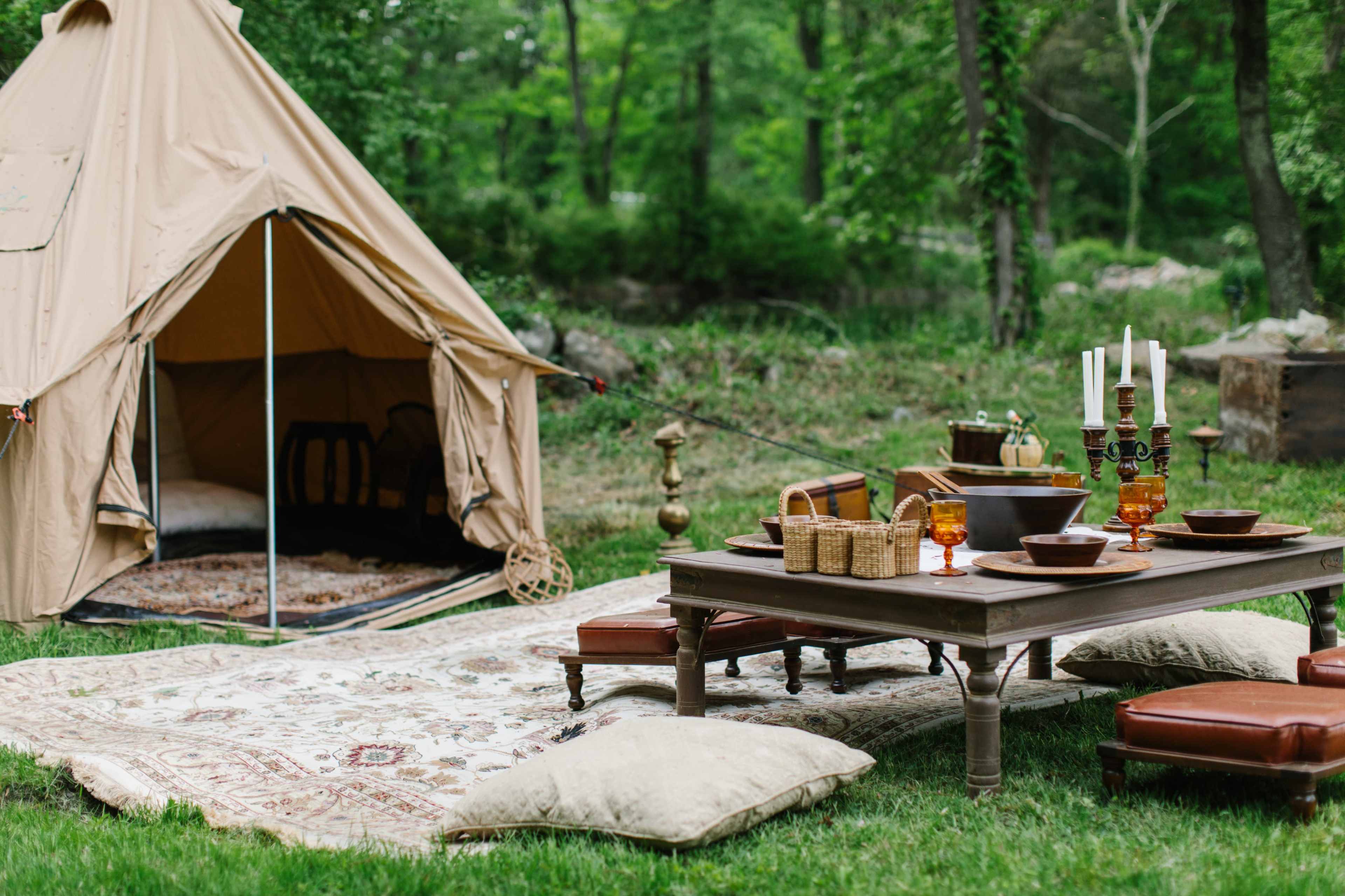 A tan canvas tent is set up in a grassy area, accompanied by a wooden table adorned with dishes and candles, surrounded by cushions and a decorative rug.