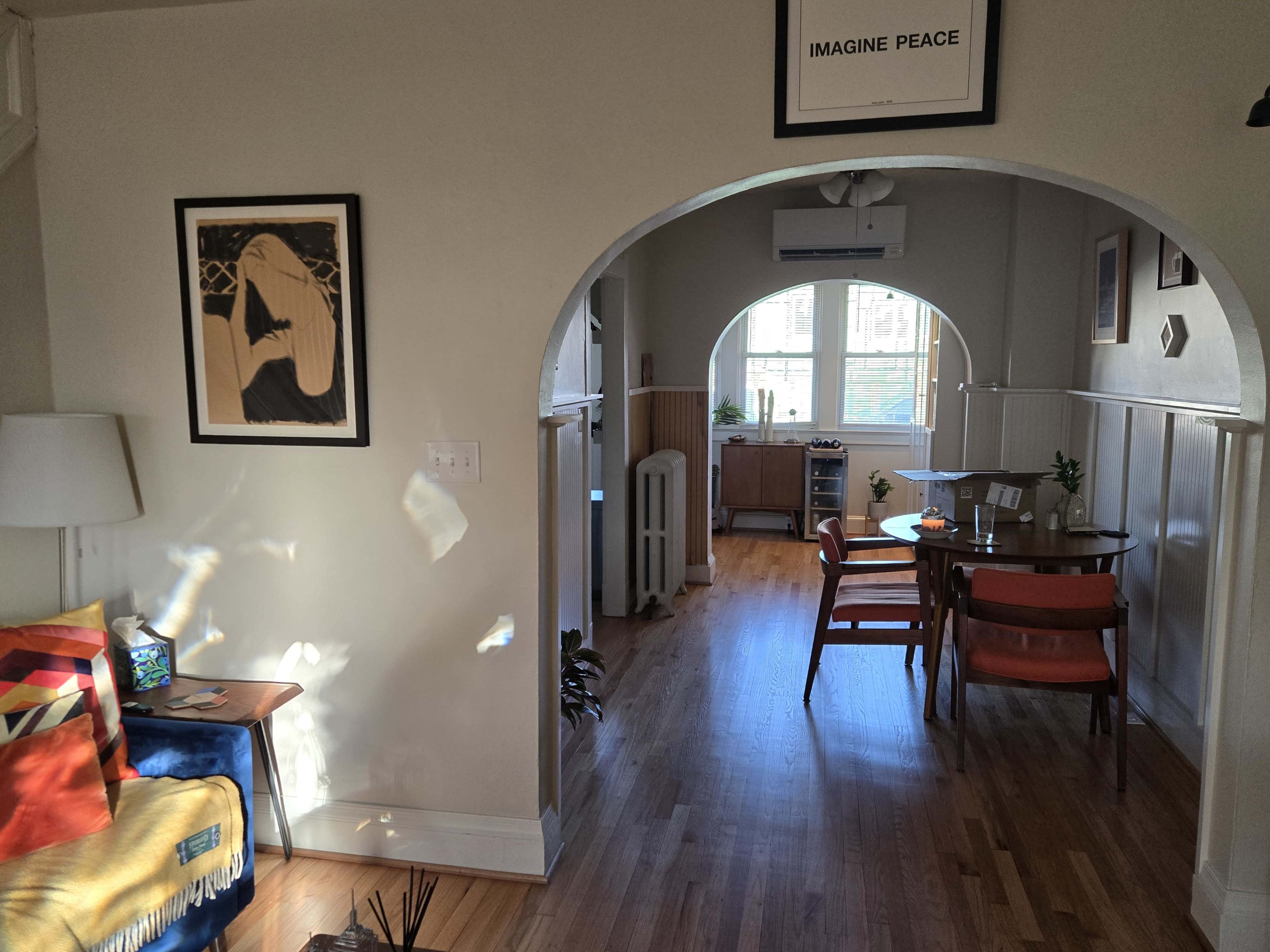 The image shows an interior of a well-lit room featuring a seating area with a blue throw, a small table, and chairs, along with an arched entrance leading to another space with a window.