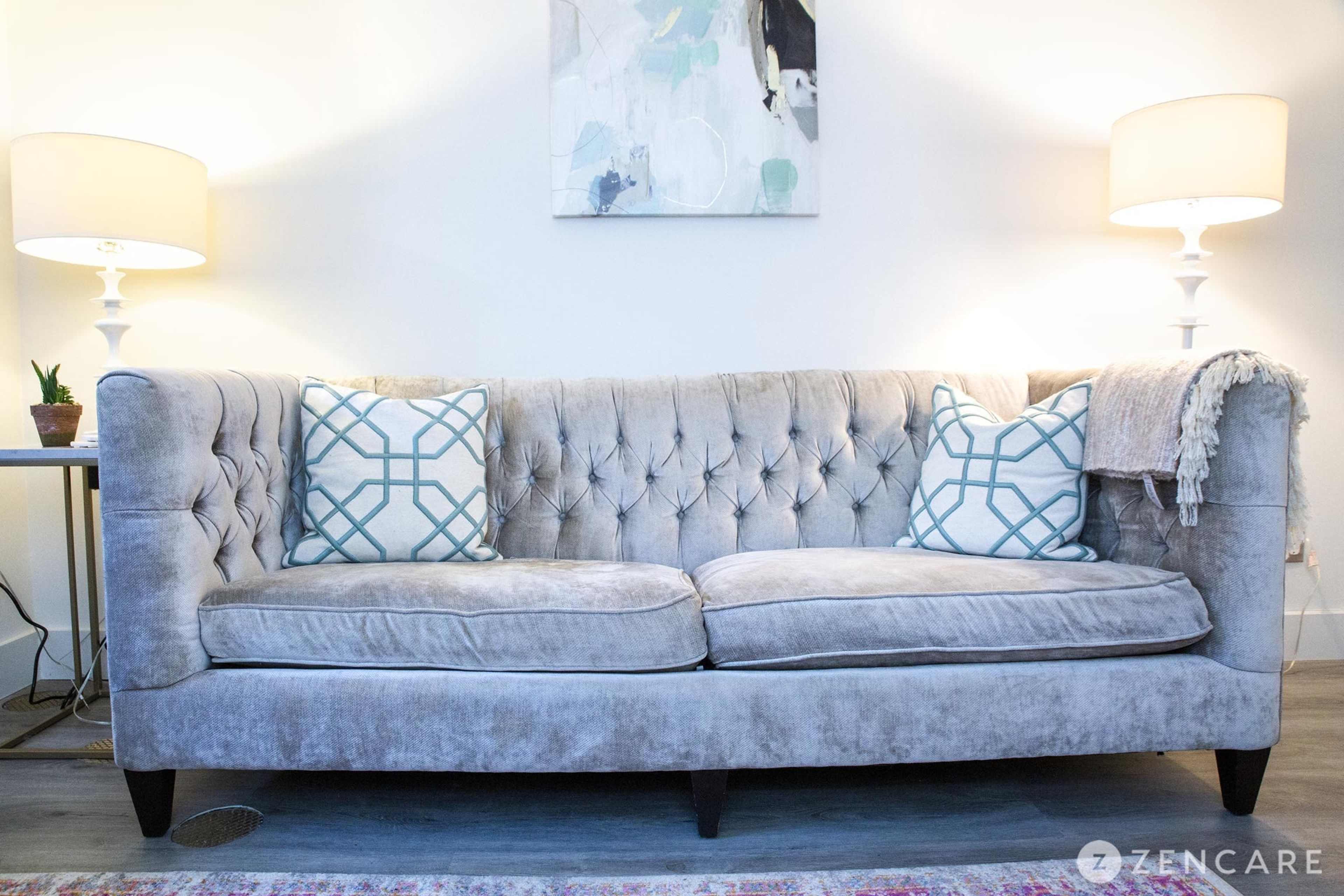 The image shows a gray tufted sofa with two decorative pillows and a throw blanket, positioned between two lamps on either side.