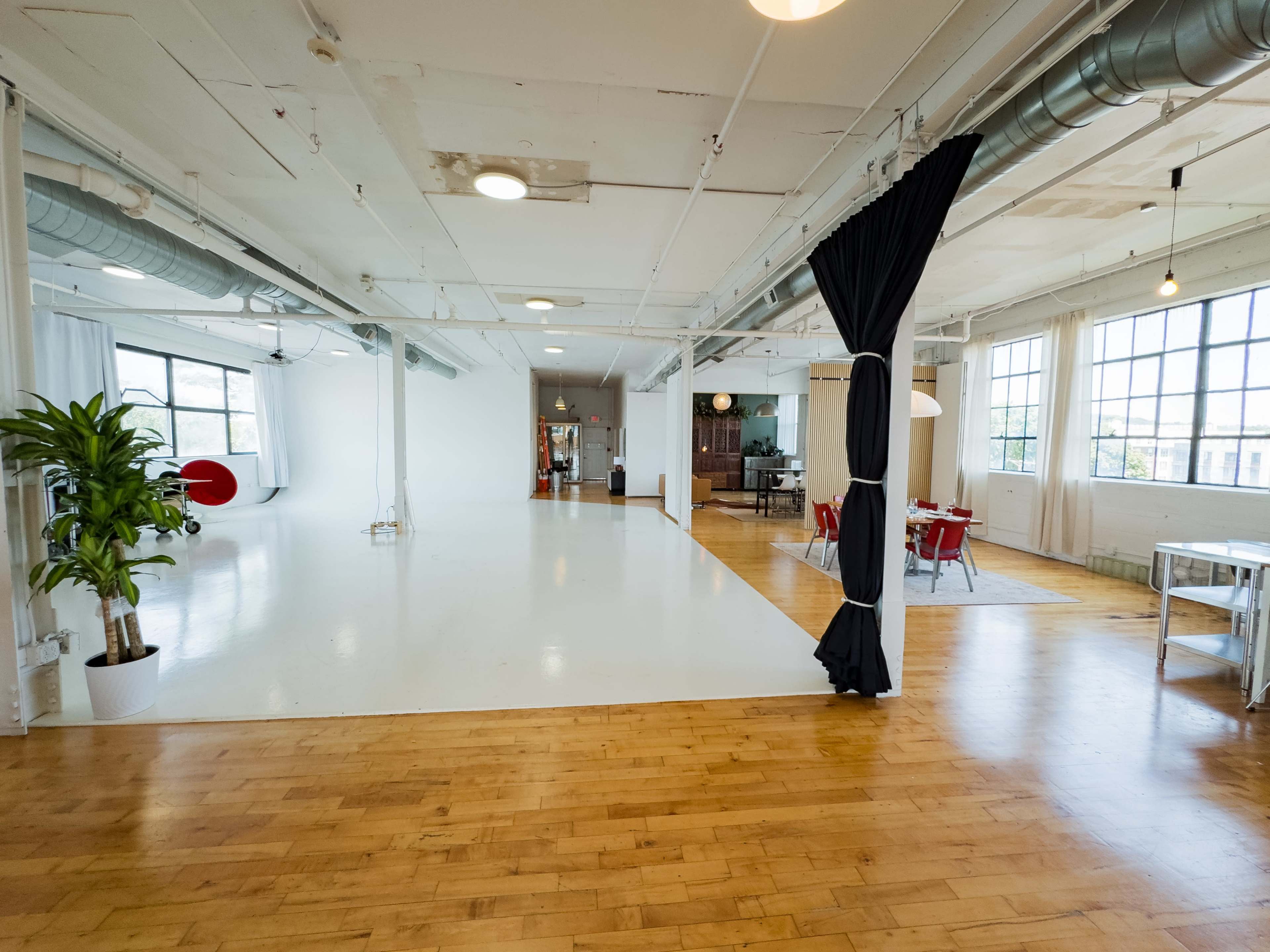 The image shows a spacious, modern industrial-style interior with polished wooden floors, large windows, and distinct areas for different activities, including a dining space and a lounge area.