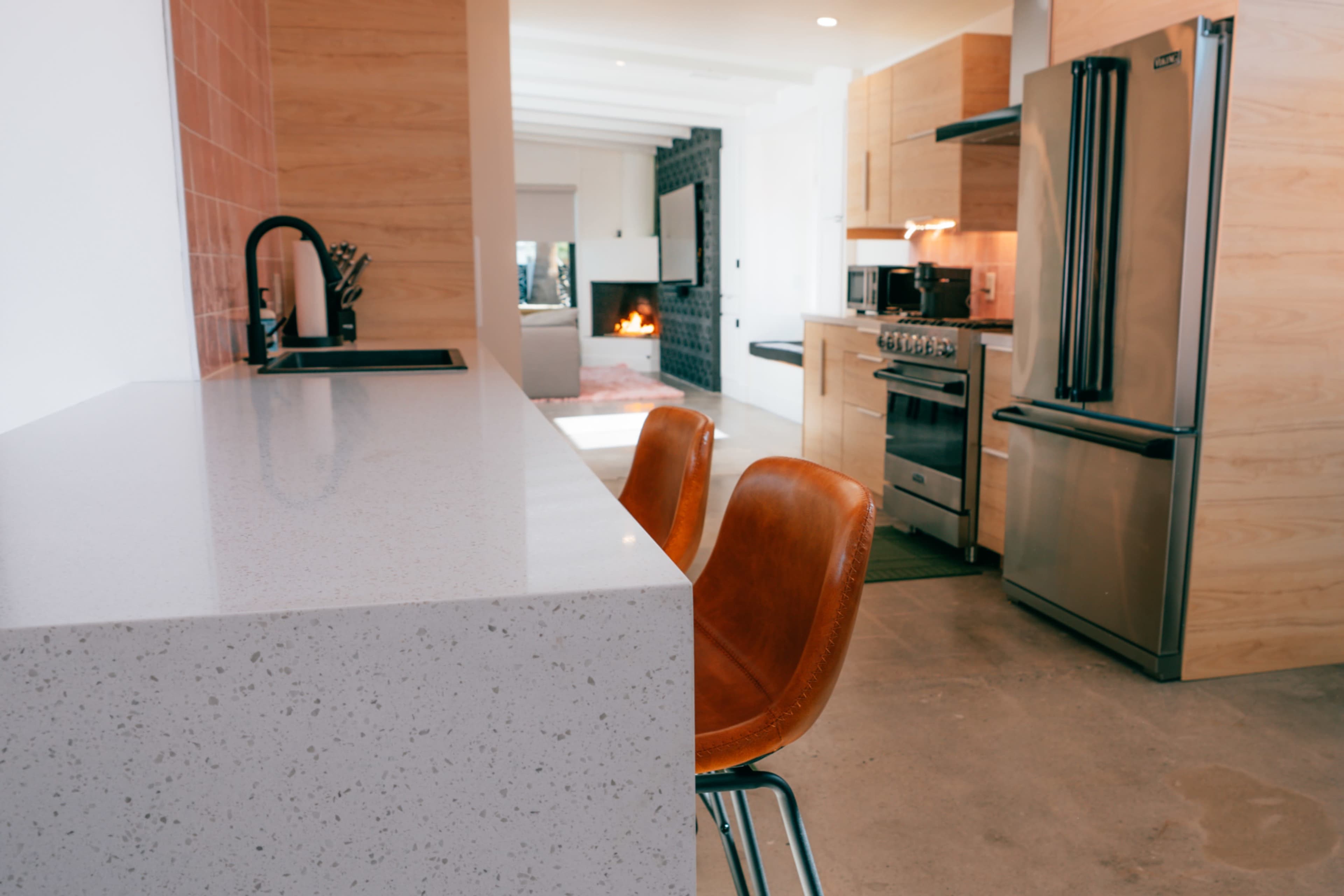 The image shows a modern kitchen with a quartz countertop and two brown leather stools, featuring stainless steel appliances and a cozy living area in the background.