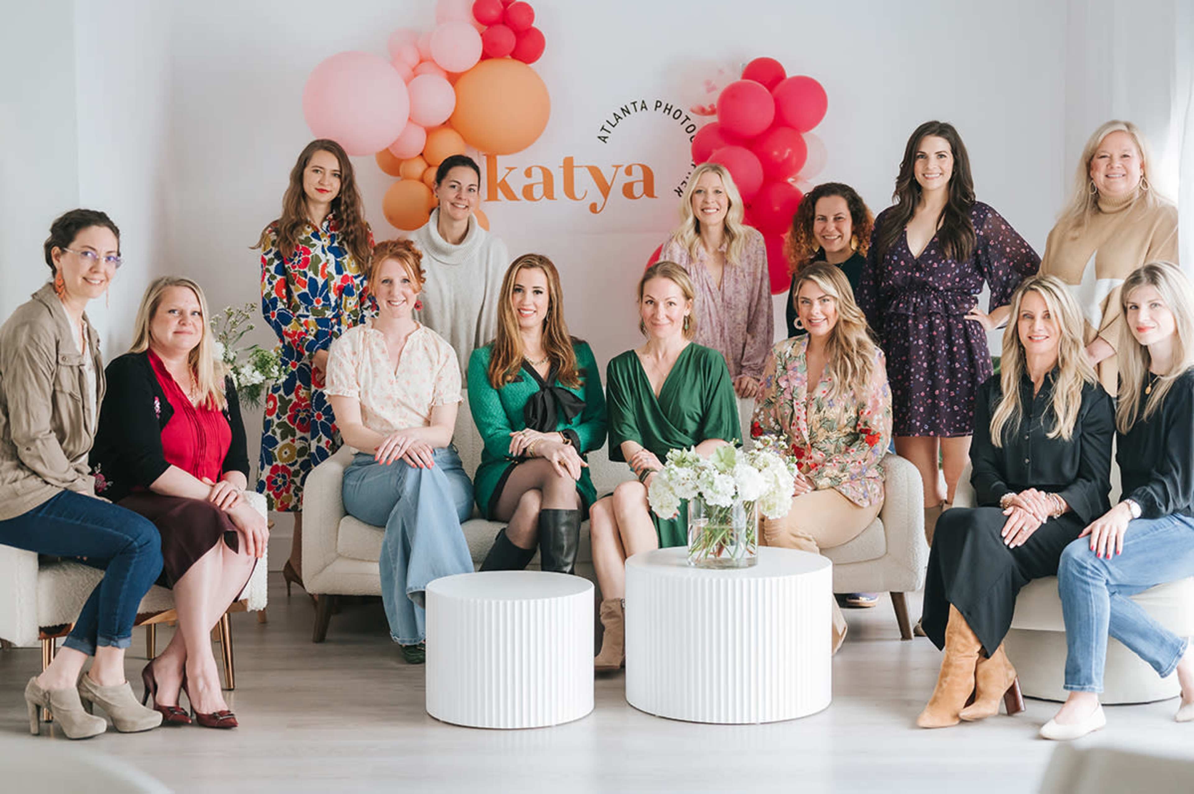 A group of women are seated together in a bright room decorated with colorful balloons, with a sign reading “katya” on the wall.