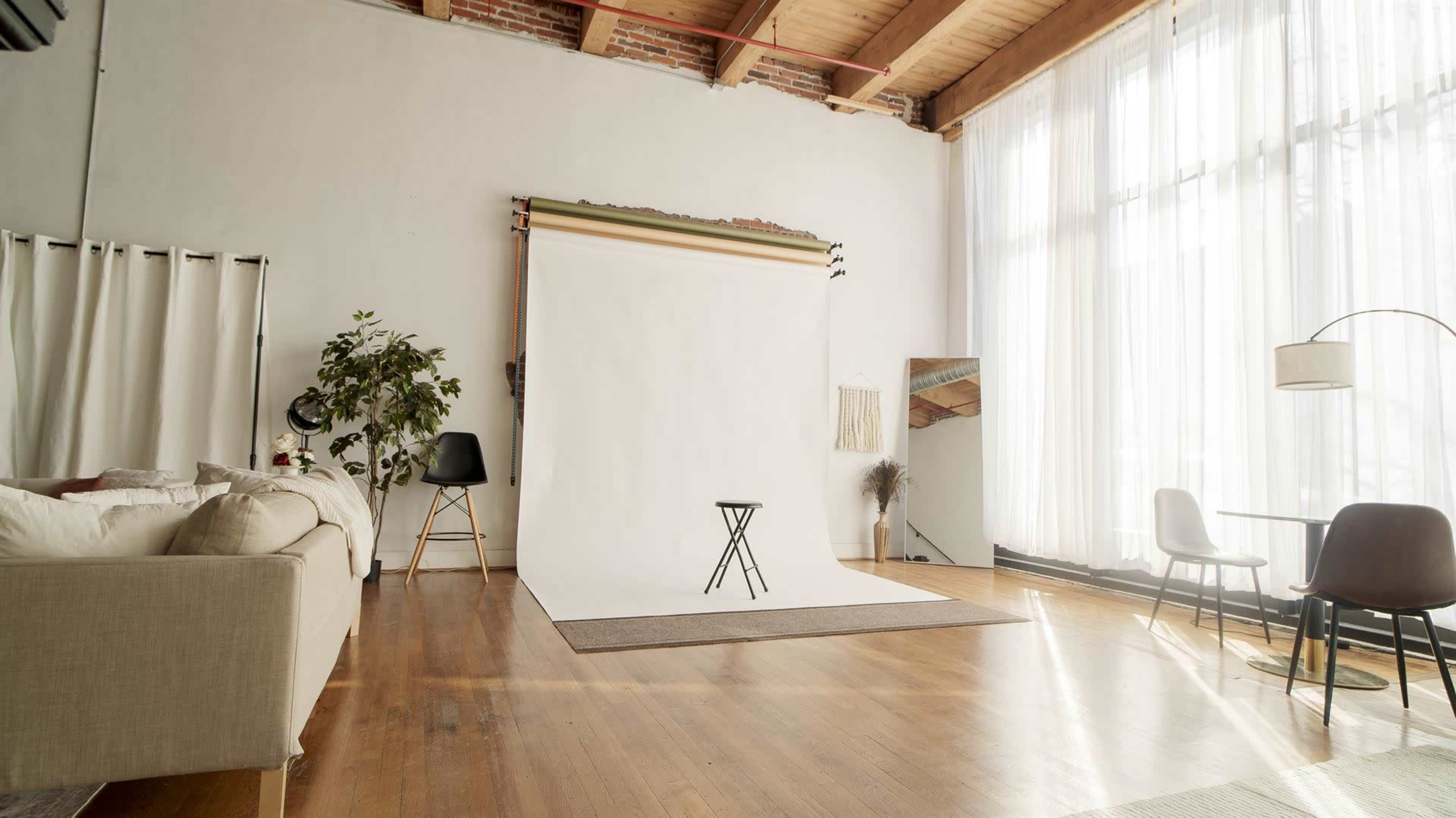The image shows a spacious photography studio with a white backdrop, a small black stool, large windows, and a mix of modern furniture.