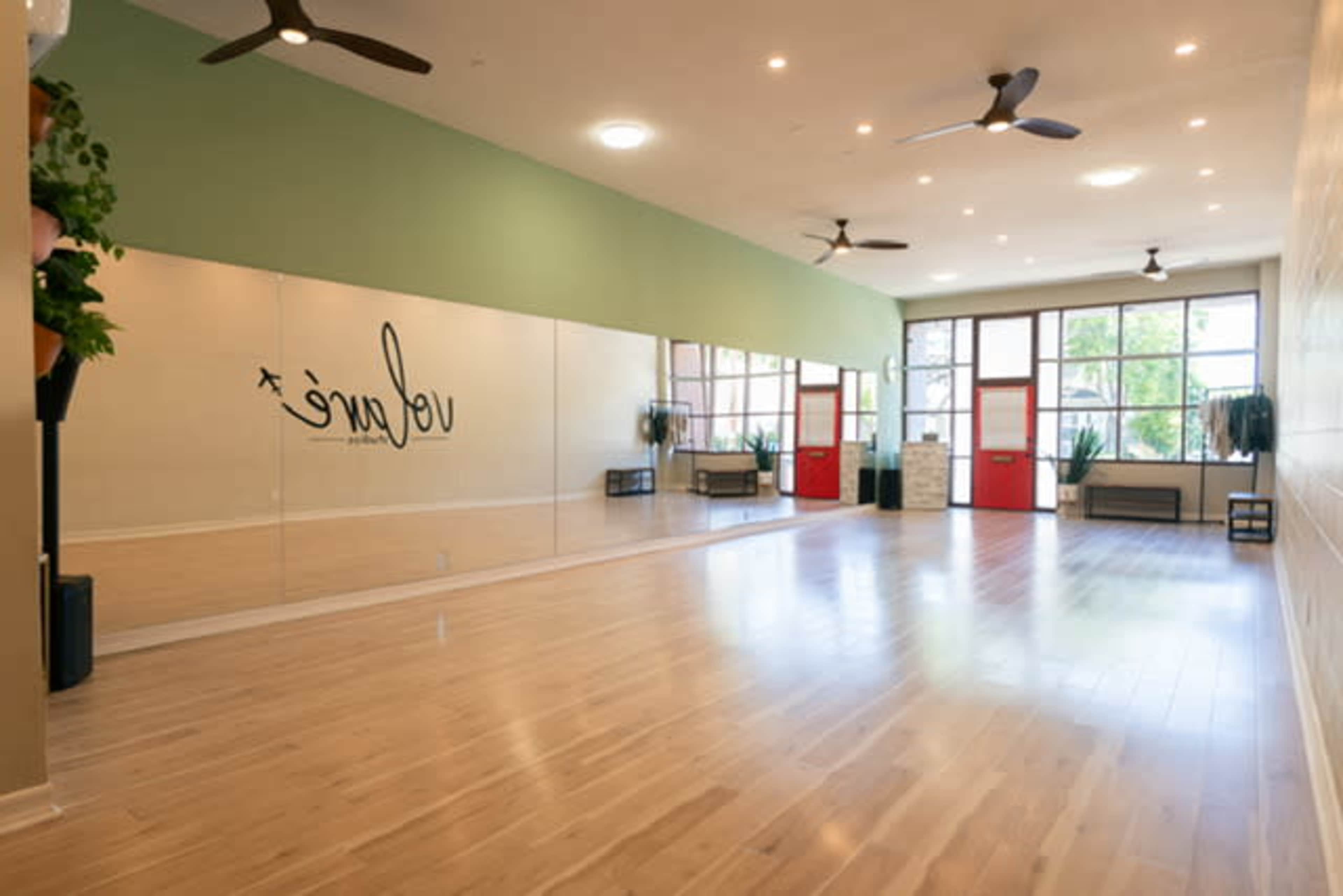 The image shows a bright and spacious studio with wooden flooring, large mirrors on one wall, and red doors leading to an outdoor area.