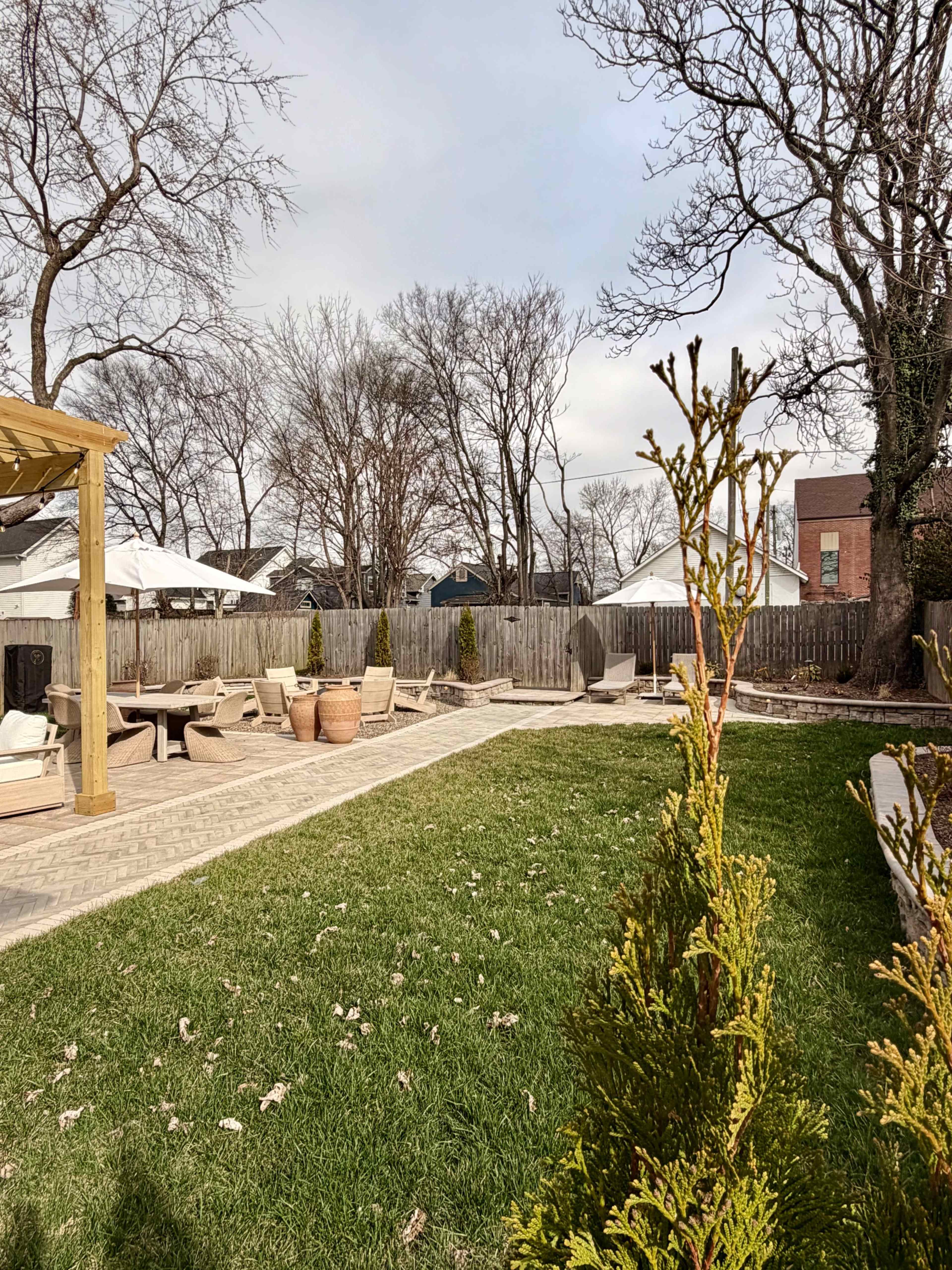 The image shows a backyard with a stone patio, garden furniture, and a grassy area bordered by trees and wooden fencing.