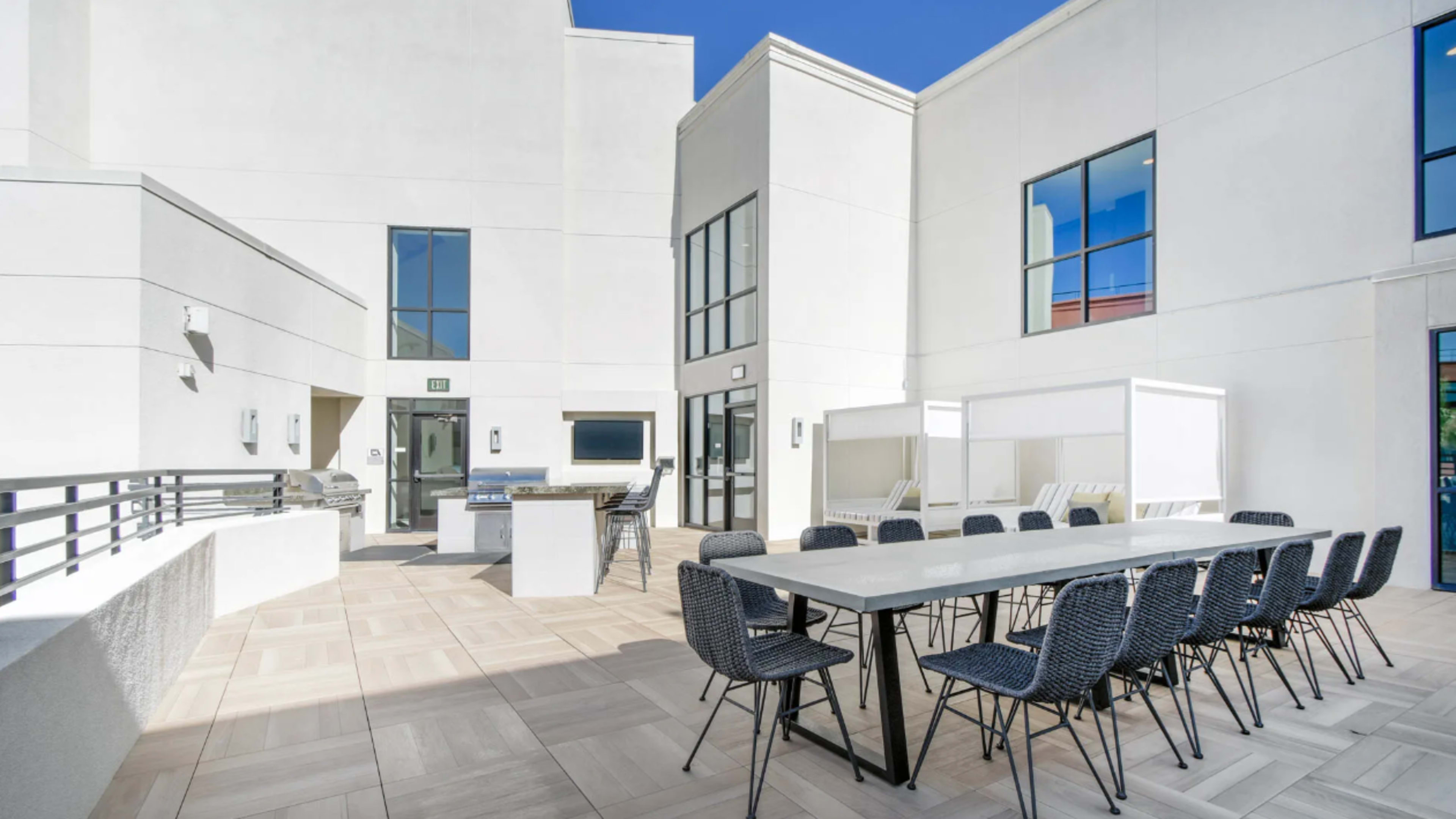 A spacious outdoor patio area with a large dining table surrounded by chairs, a grill station, and white cabanas against a backdrop of modern white buildings.