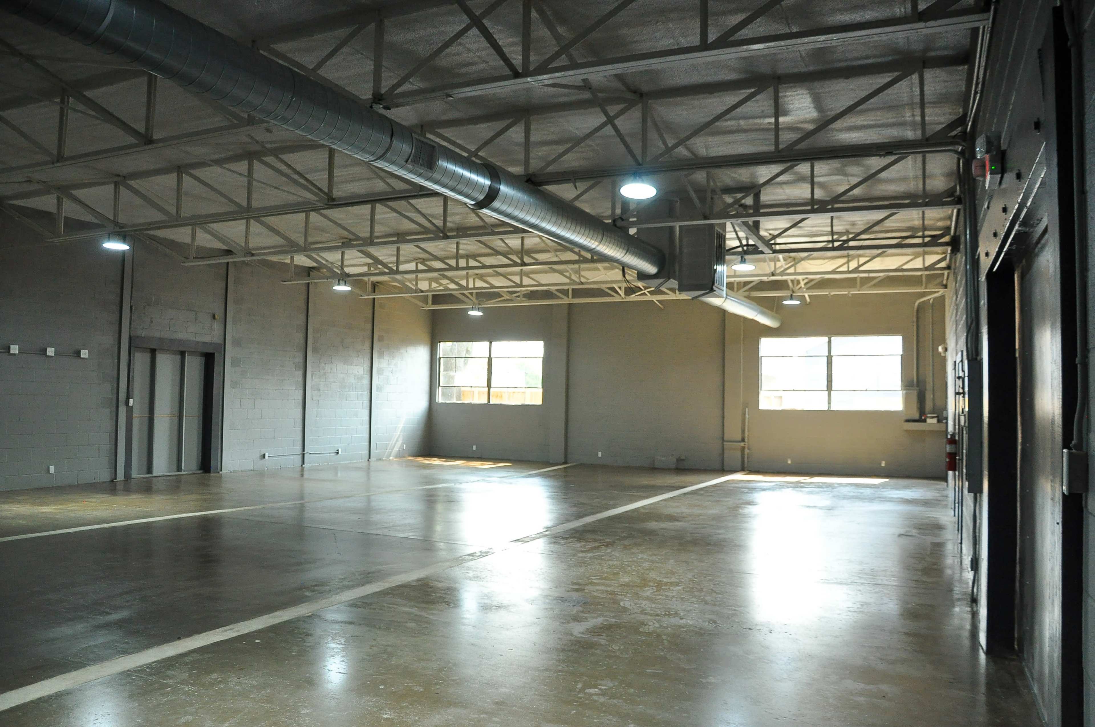 A spacious, empty industrial warehouse with concrete flooring, high ceilings, and large windows providing natural light.