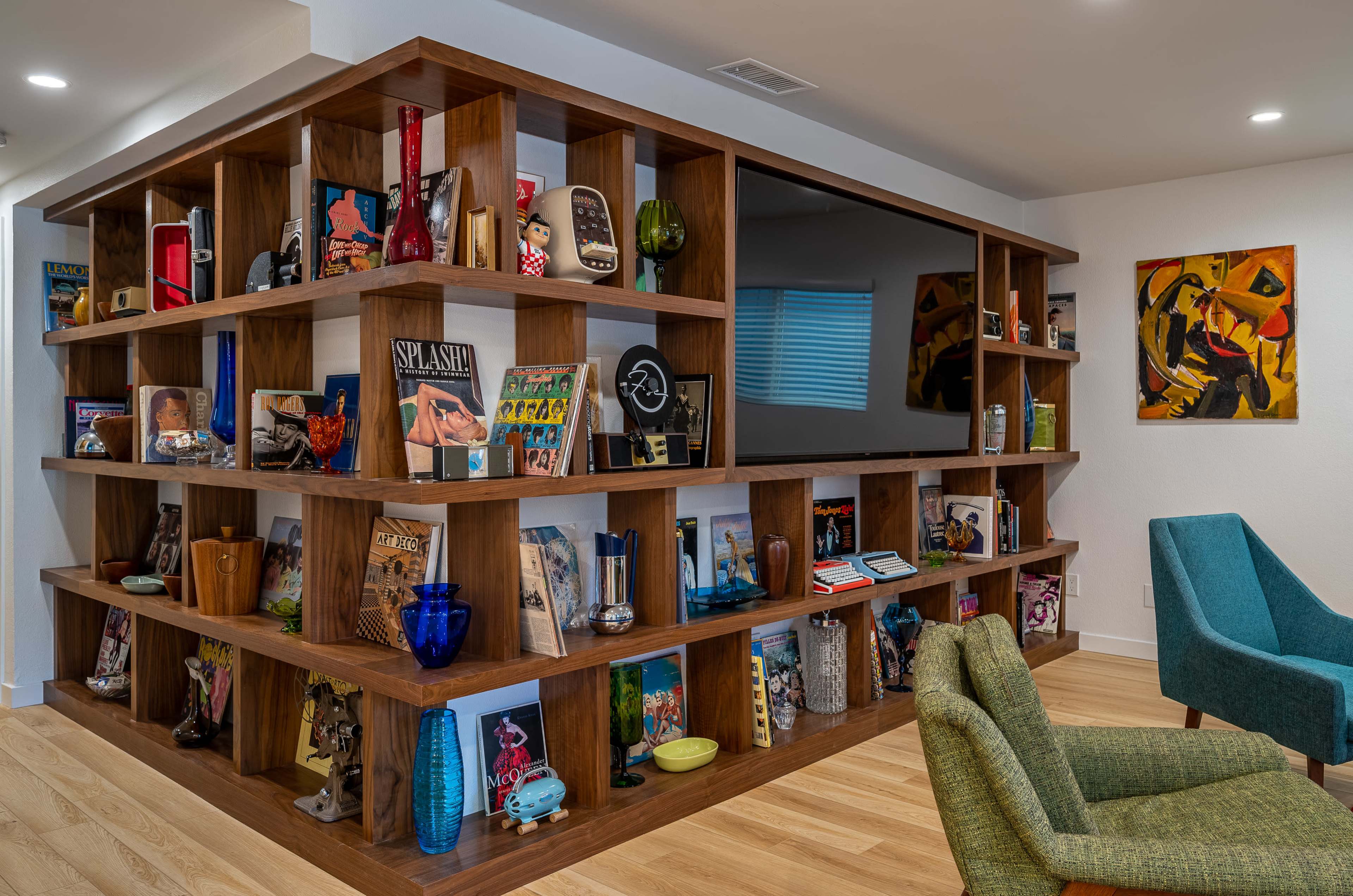 A modern room features a wooden shelf filled with books, magazines, and decorative items, alongside two colorful chairs.