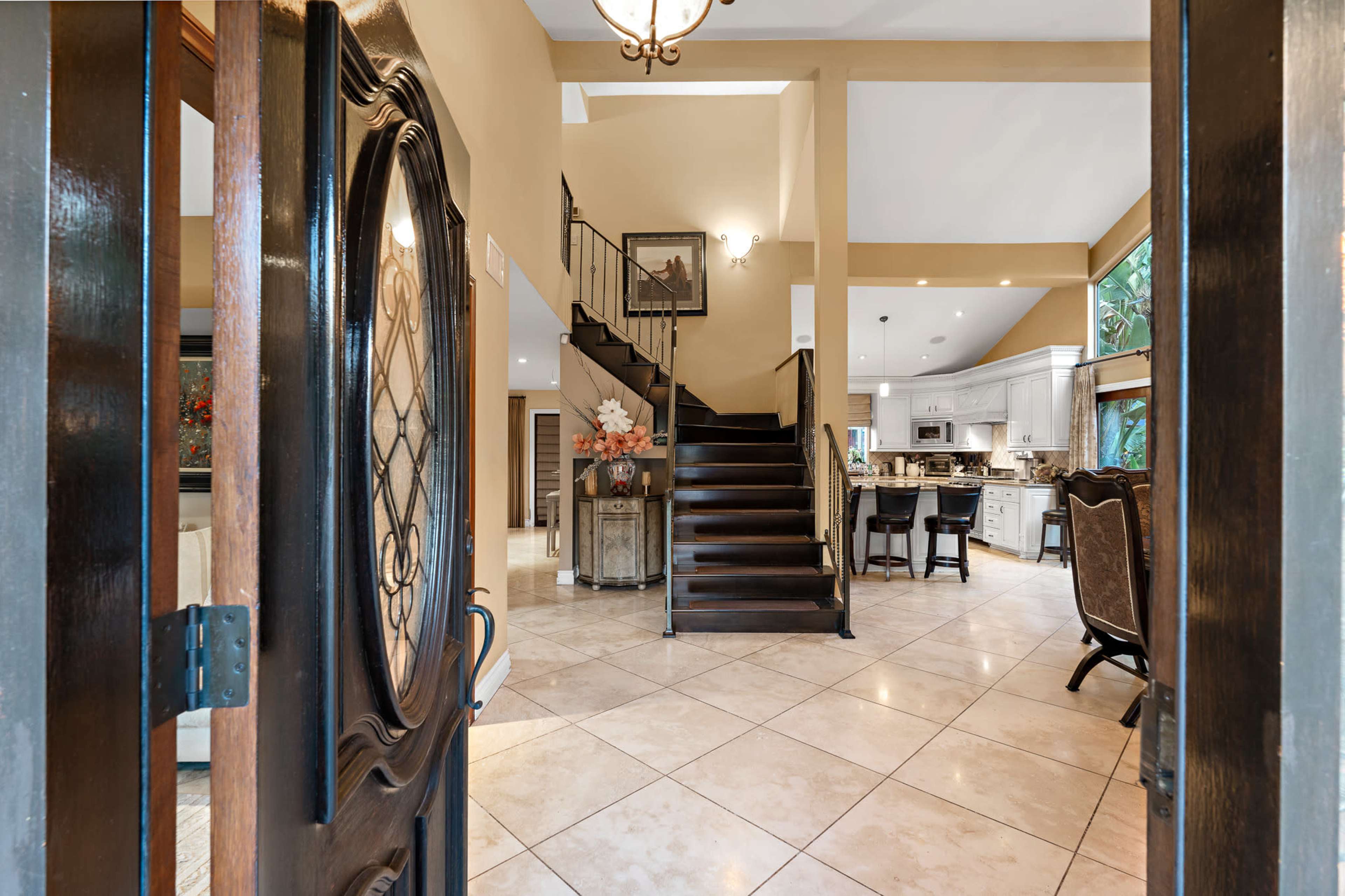 The image shows the entrance to a home, revealing a foyer with a staircase leading to an upper level, tiled flooring, and an open view into a kitchen and dining area.