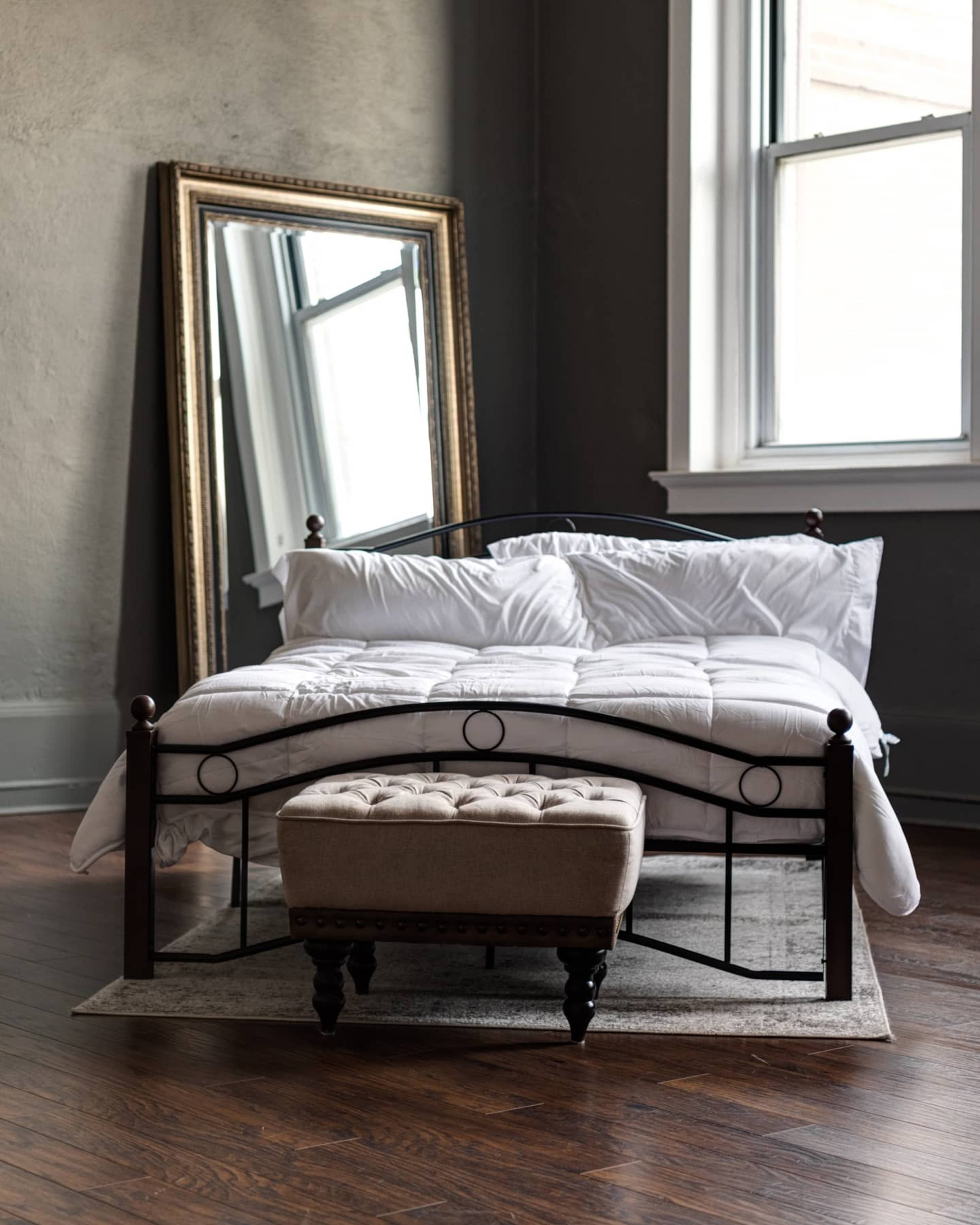A metal bed with white bedding sits in a room featuring a large mirror and a tufted stool on a rug.