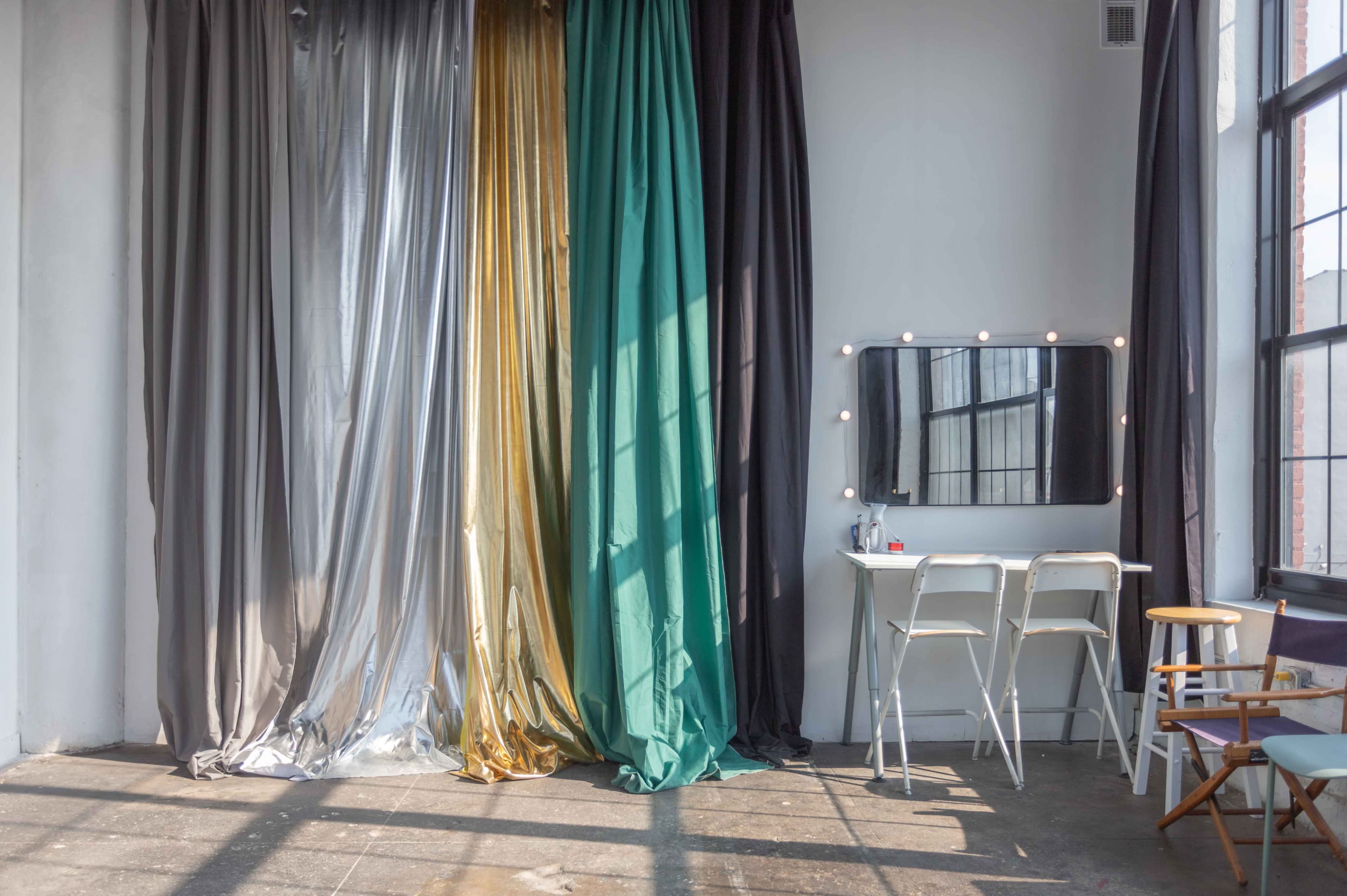 A bright room featuring colorful curtains in silver, gold, green, and black, along with a simple table and chairs beside a mirror with string lights.