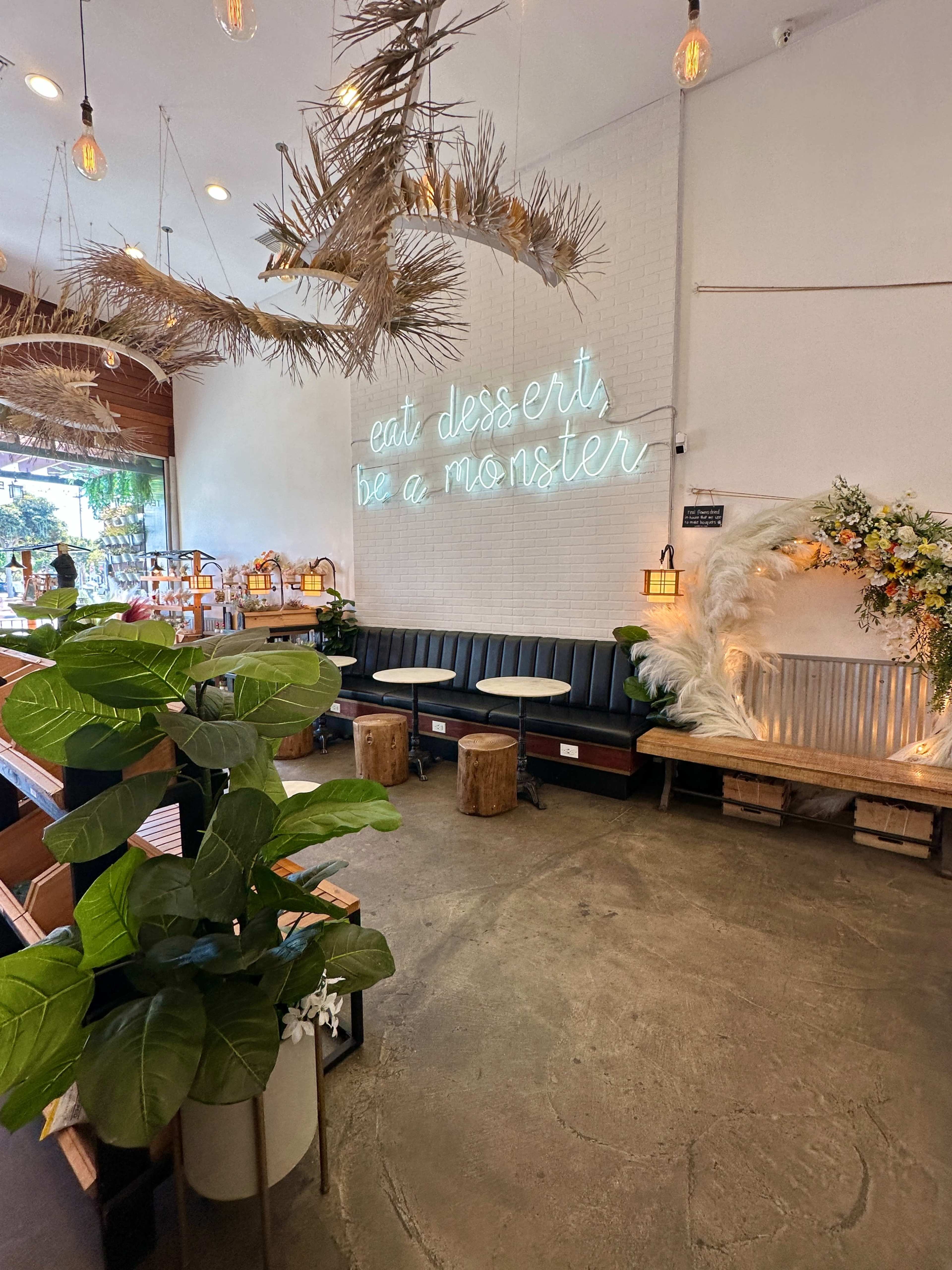 The interior features a cozy seating area with wooden benches, pendant lights, tropical plants, and a neon sign that reads, "eat desserts, be a monster."