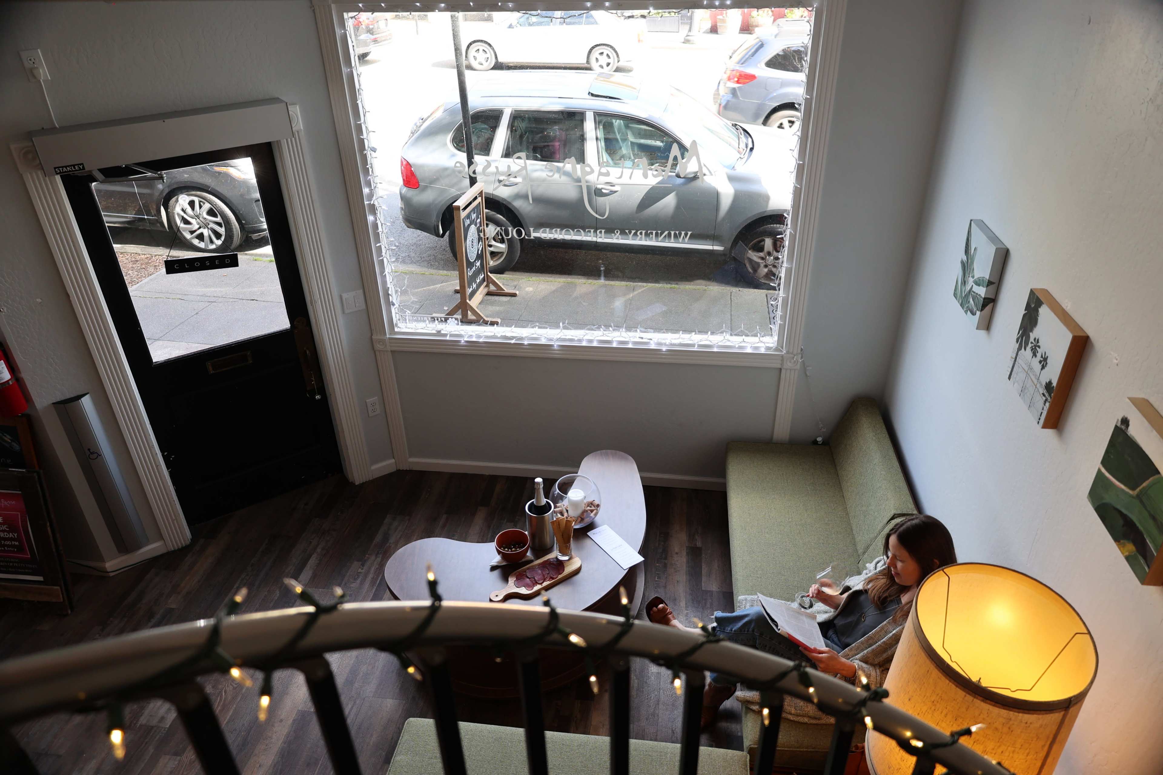A woman sits on a green couch in a cafe, with a view of parked cars outside through a large window.