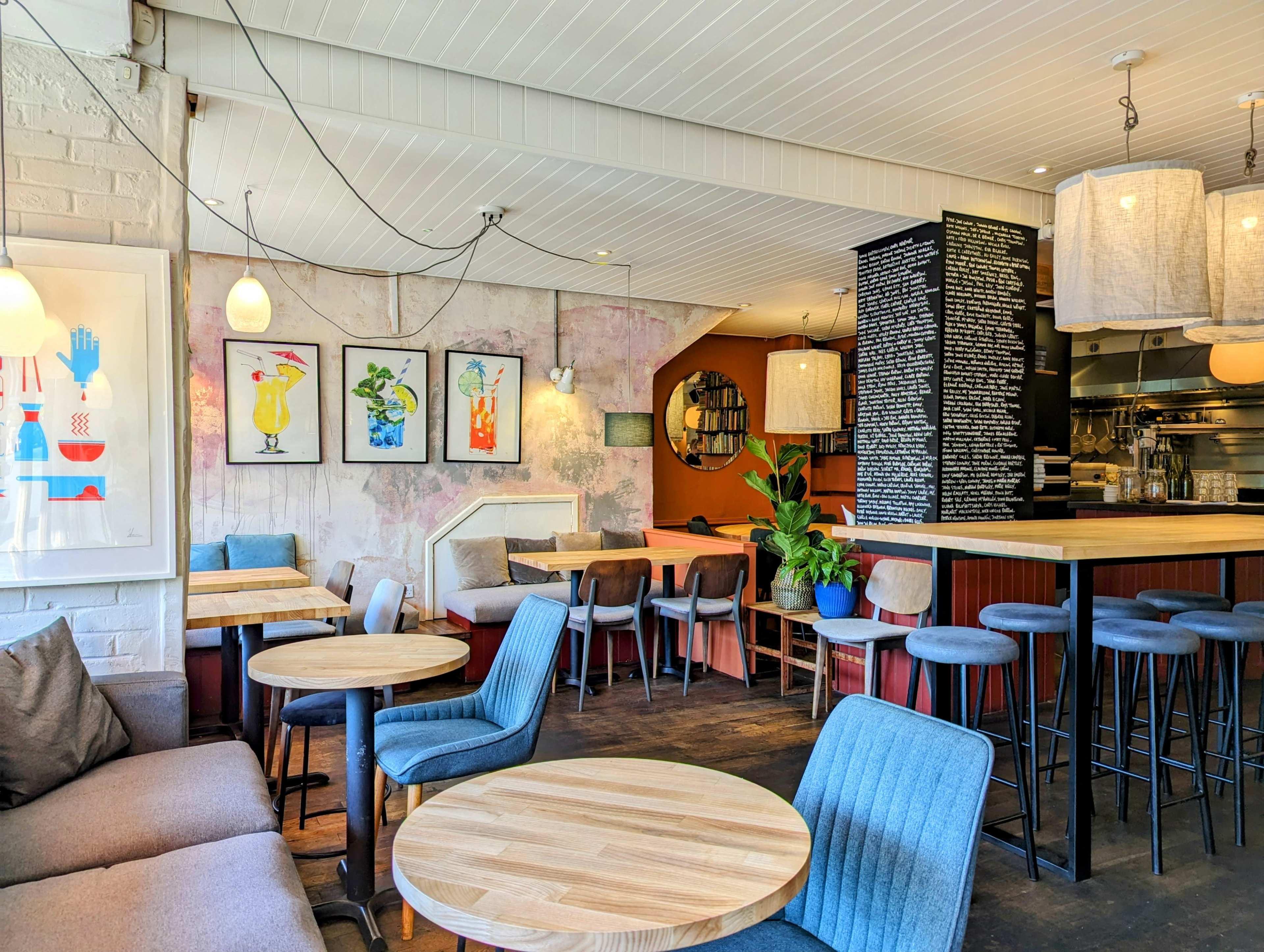 The image shows a cozy café interior with wooden tables, blue and gray seating, colorful artwork on the walls, and a bar area in the background.