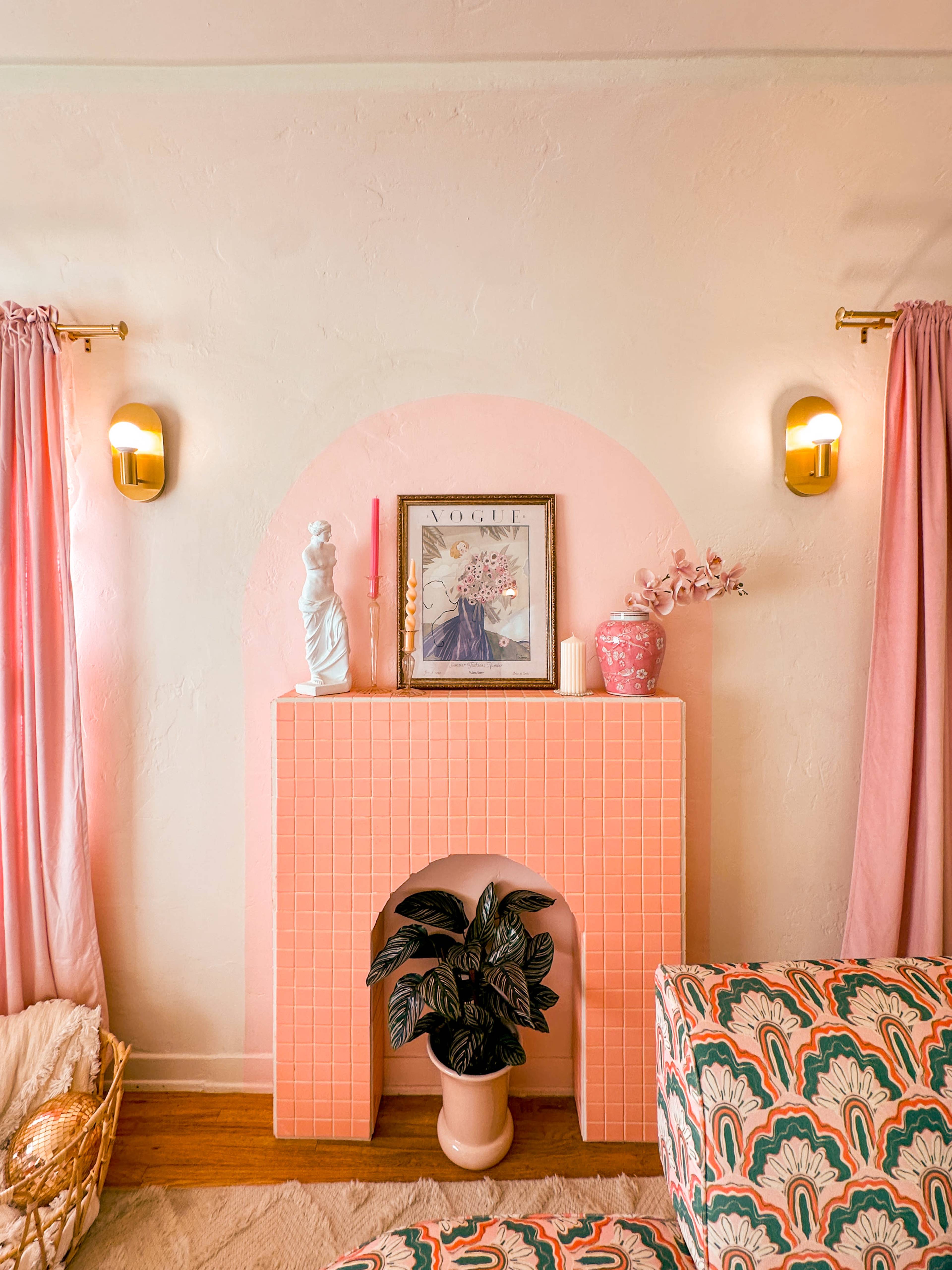 The image shows a pastel-colored living room featuring a pink-tiled fireplace adorned with decorative items, framed art, and plants, complemented by soft pink curtains and a patterned sofa.
