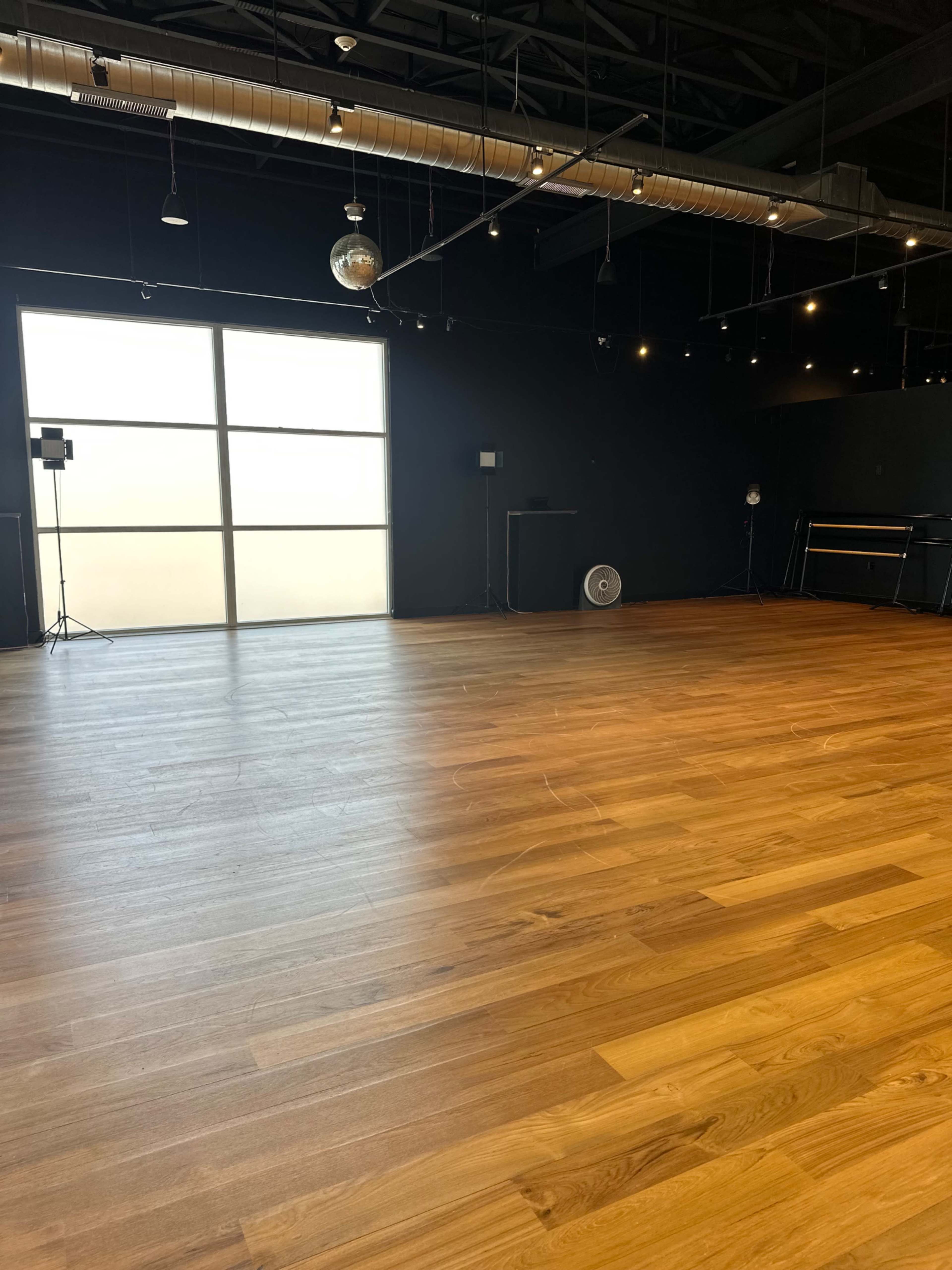 The image shows a spacious, empty dance studio with wooden flooring, large windows, and a few light fixtures.