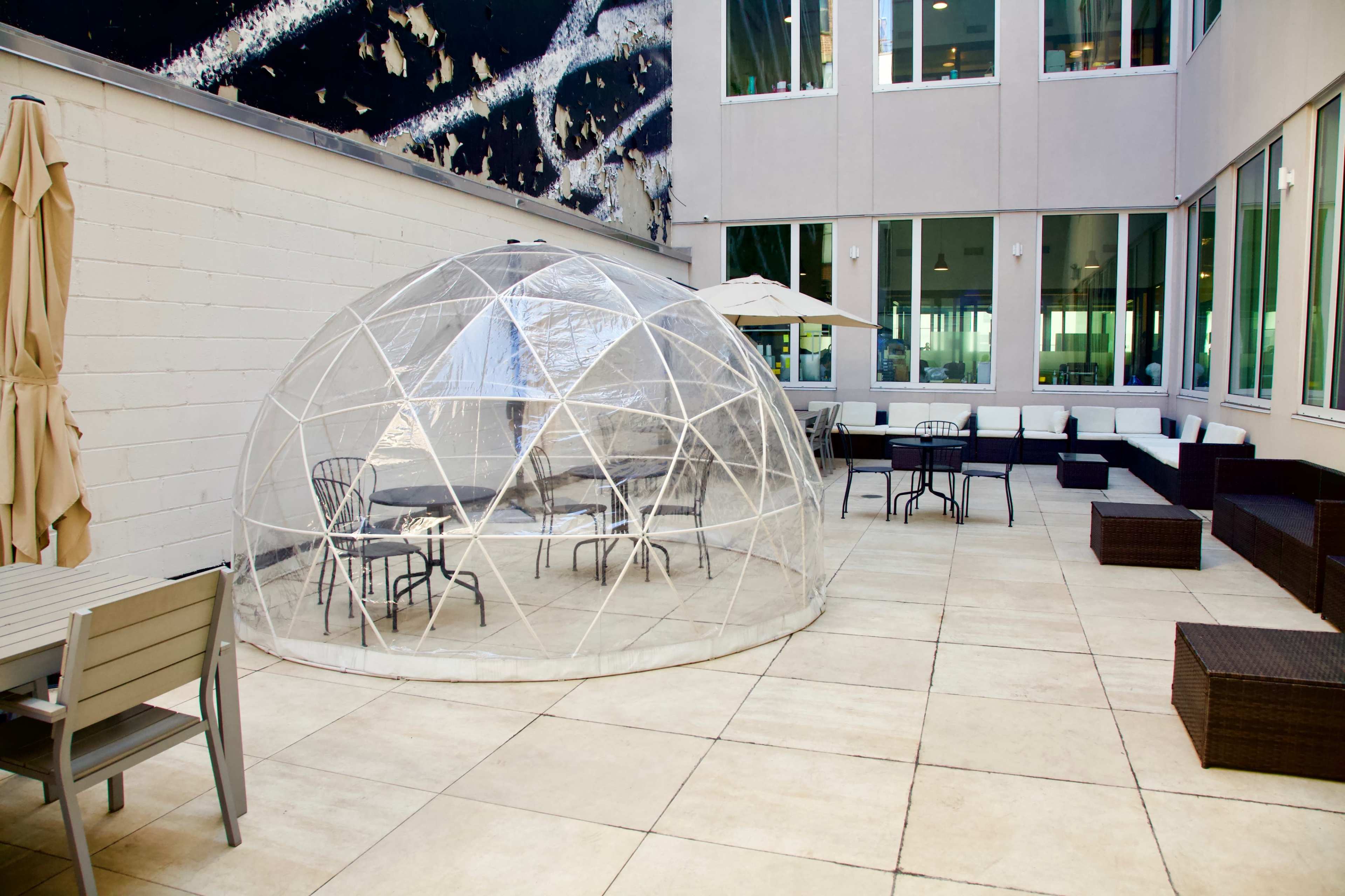 A clear geodesic dome sits on a tile patio surrounded by outdoor furniture and large windows.