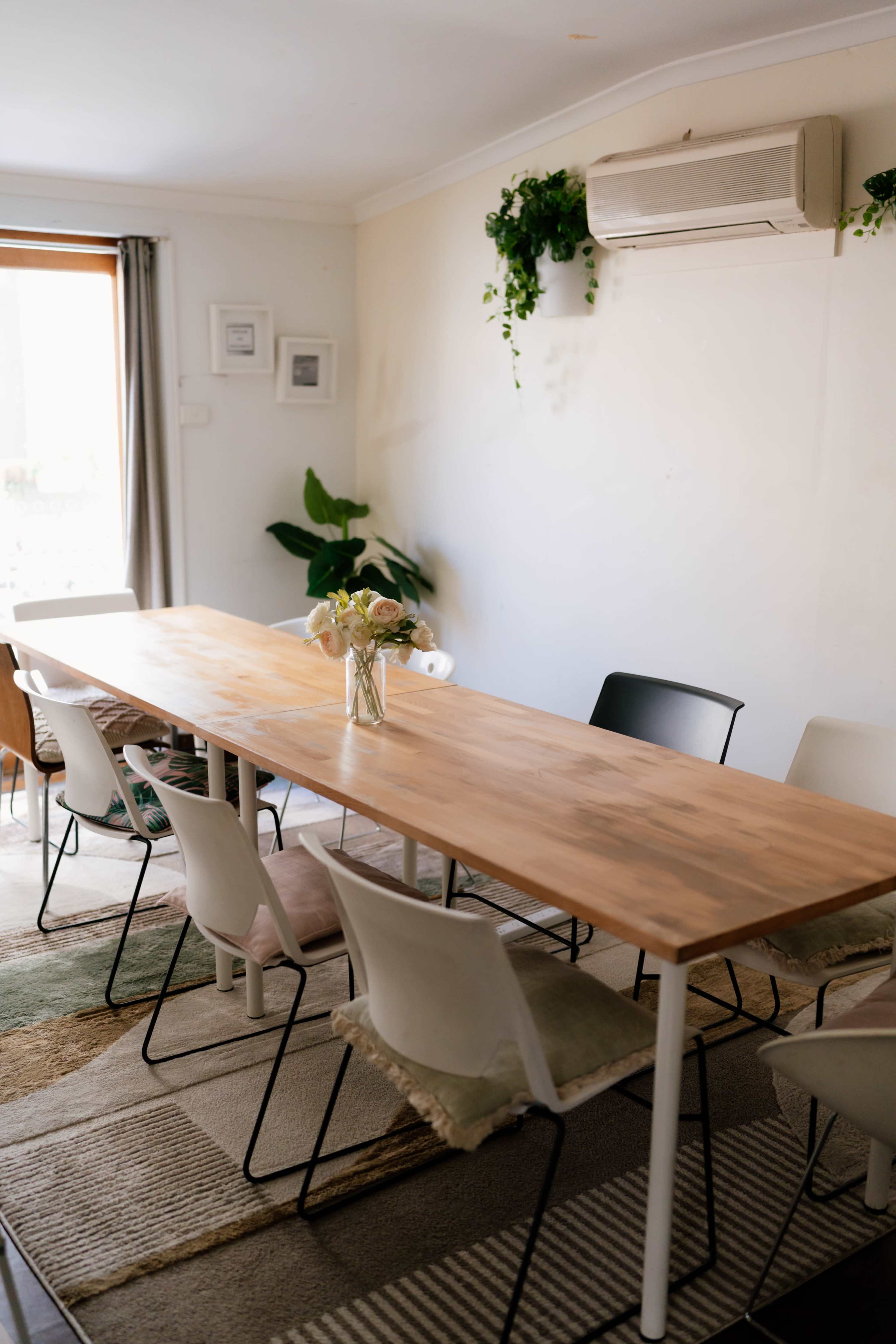 A long wooden table is set in a bright room surrounded by several white and black chairs, with a vase of flowers at the center and plants adorning the walls.
