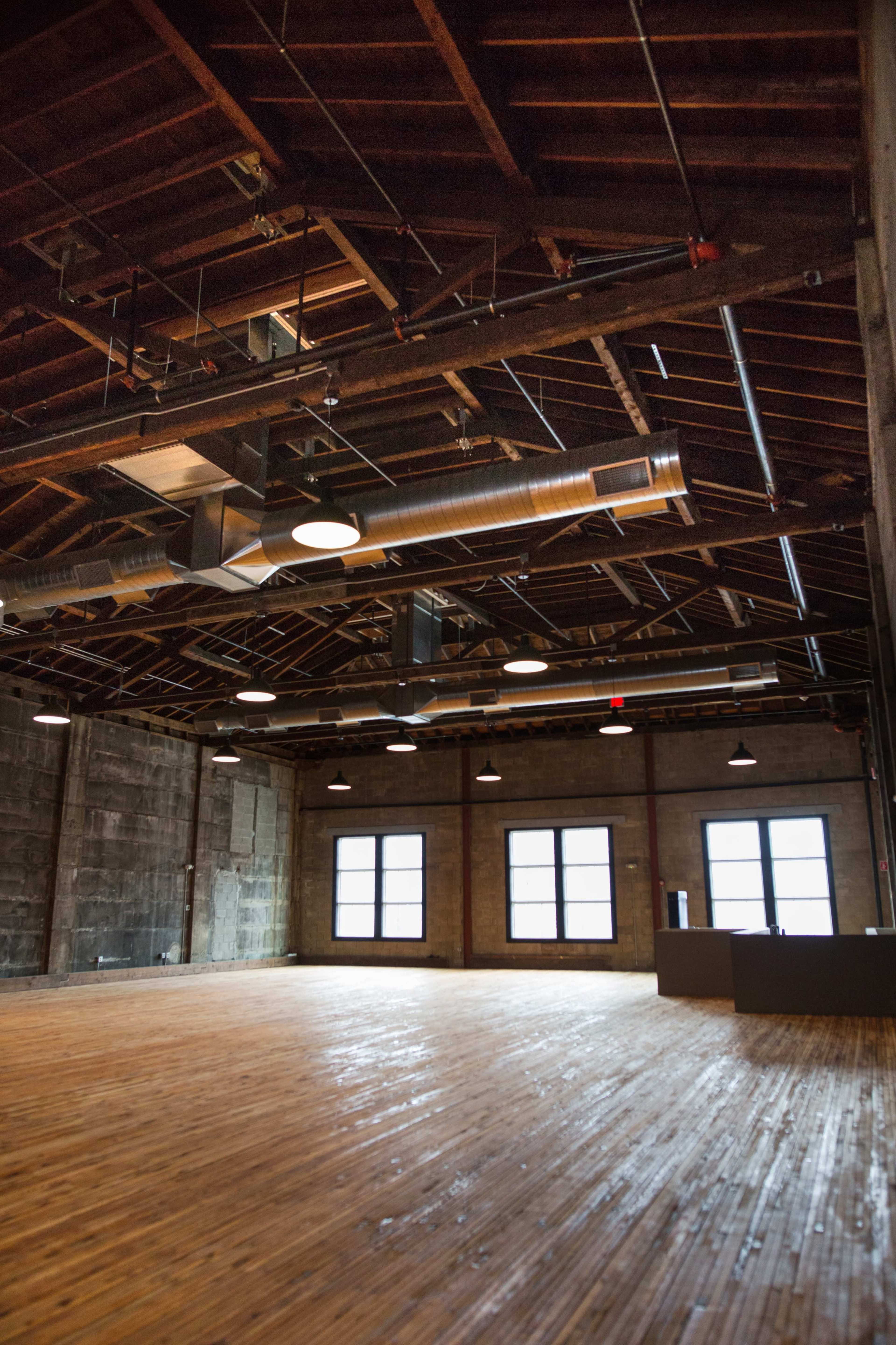 The image shows an empty industrial-style room with wooden floors, exposed beams, and large windows.
