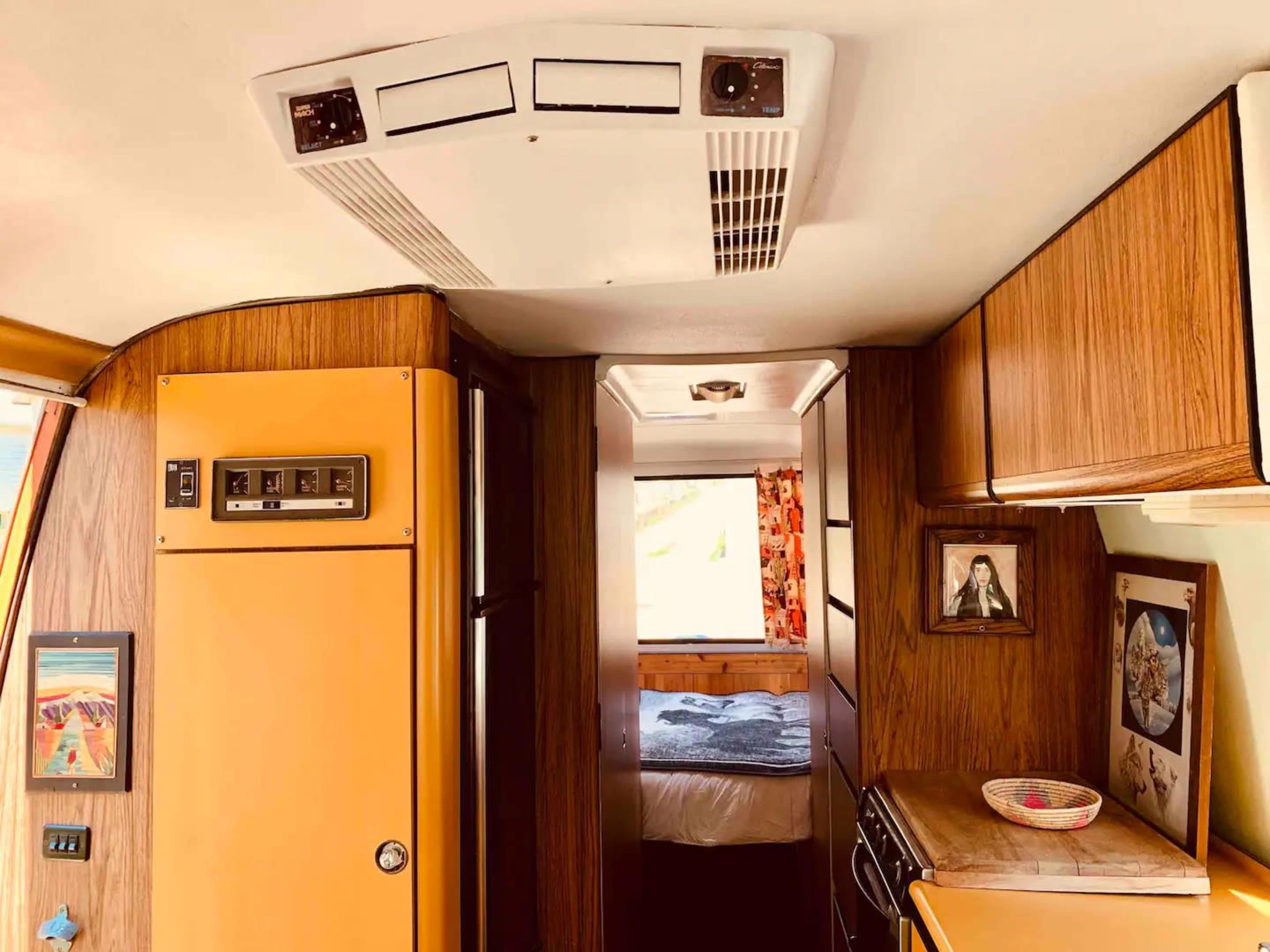 The interior of a vintage camper features wooden cabinetry, a small kitchen area with appliances, and an adjacent bedroom visible through a doorway.