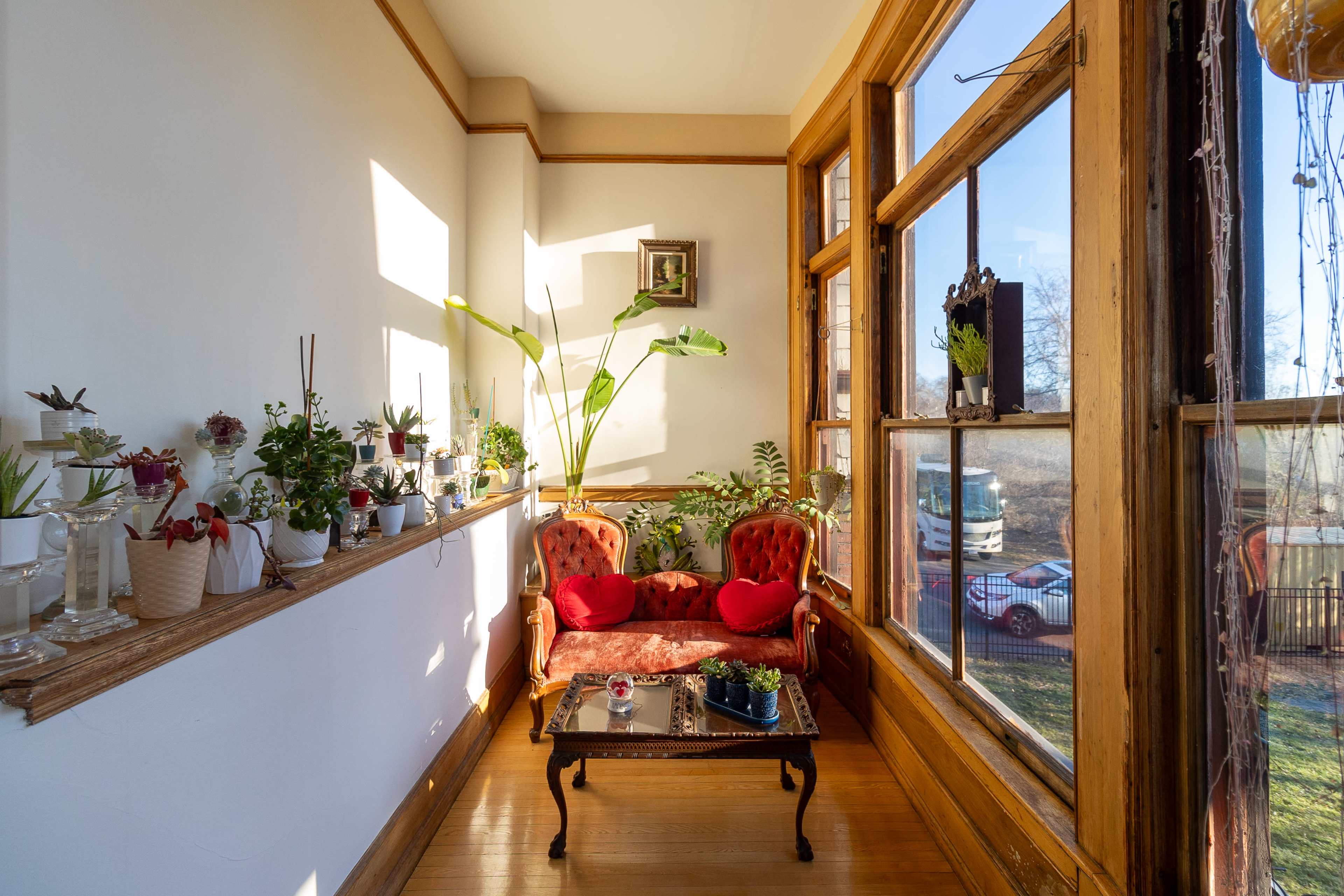 A bright sunroom features a red sofa with two cushions, a small coffee table, and various potted plants arranged near large windows.