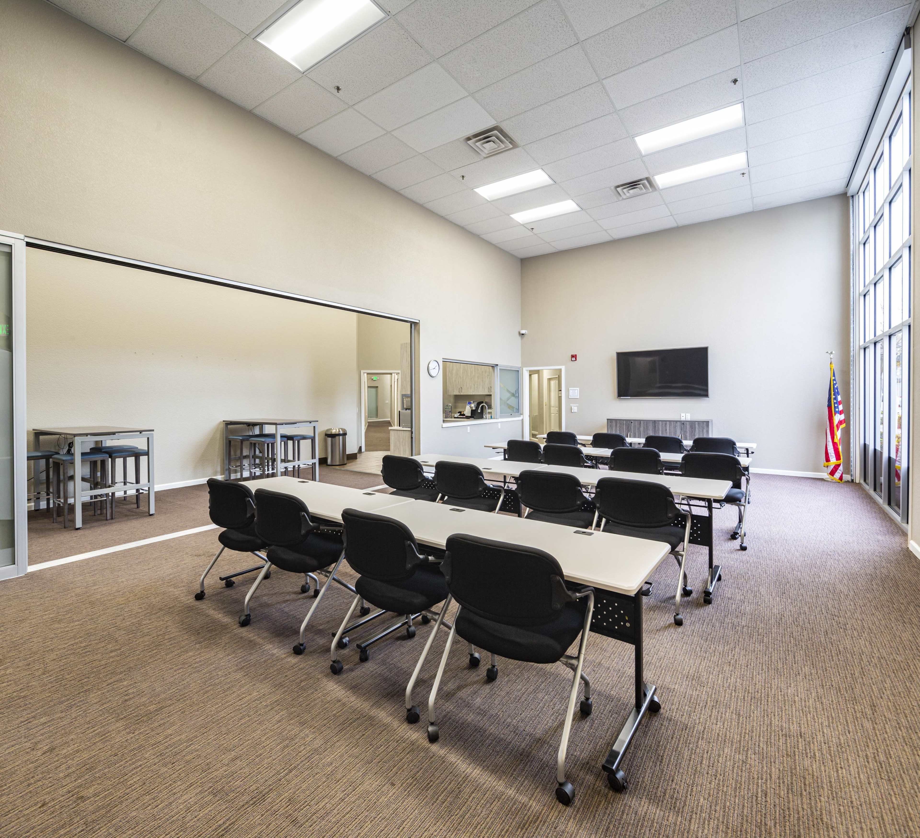 A spacious, well-lit classroom with multiple tables and chairs arranged in a grid, featuring a large wall-mounted television and an American flag in the corner.