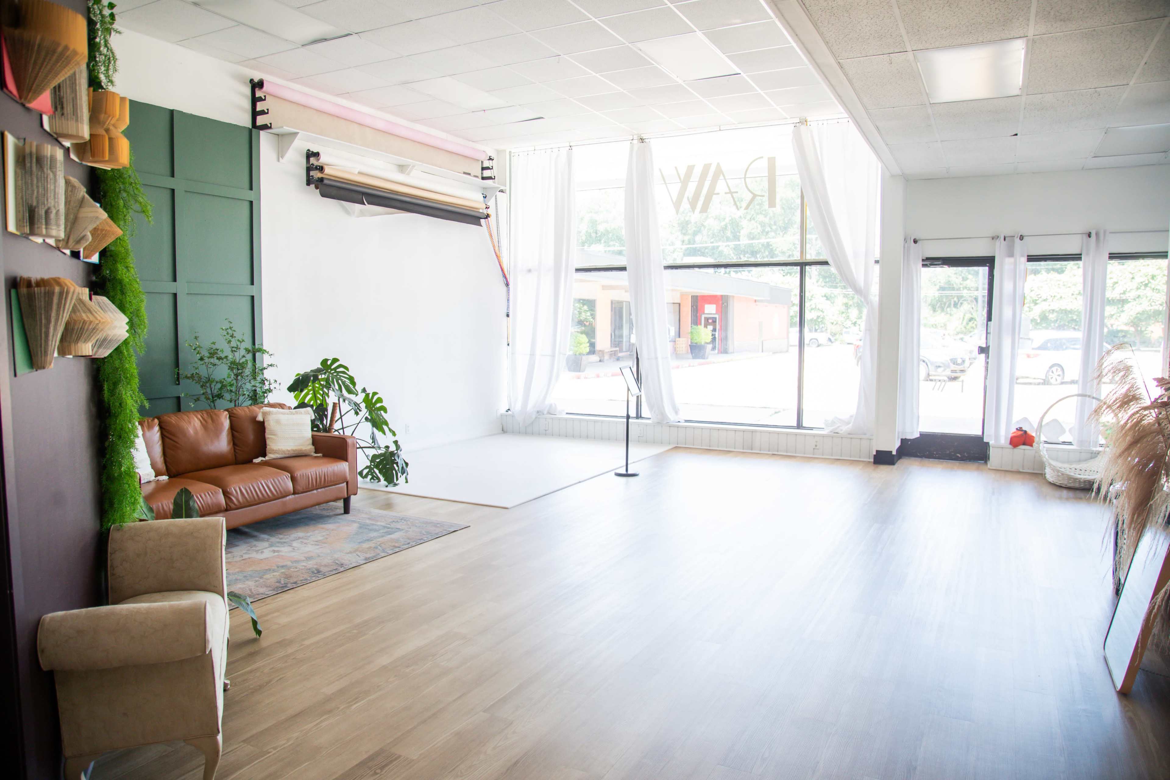 The image shows a spacious, well-lit studio with a brown leather couch, large windows, and a minimalistic decor style.