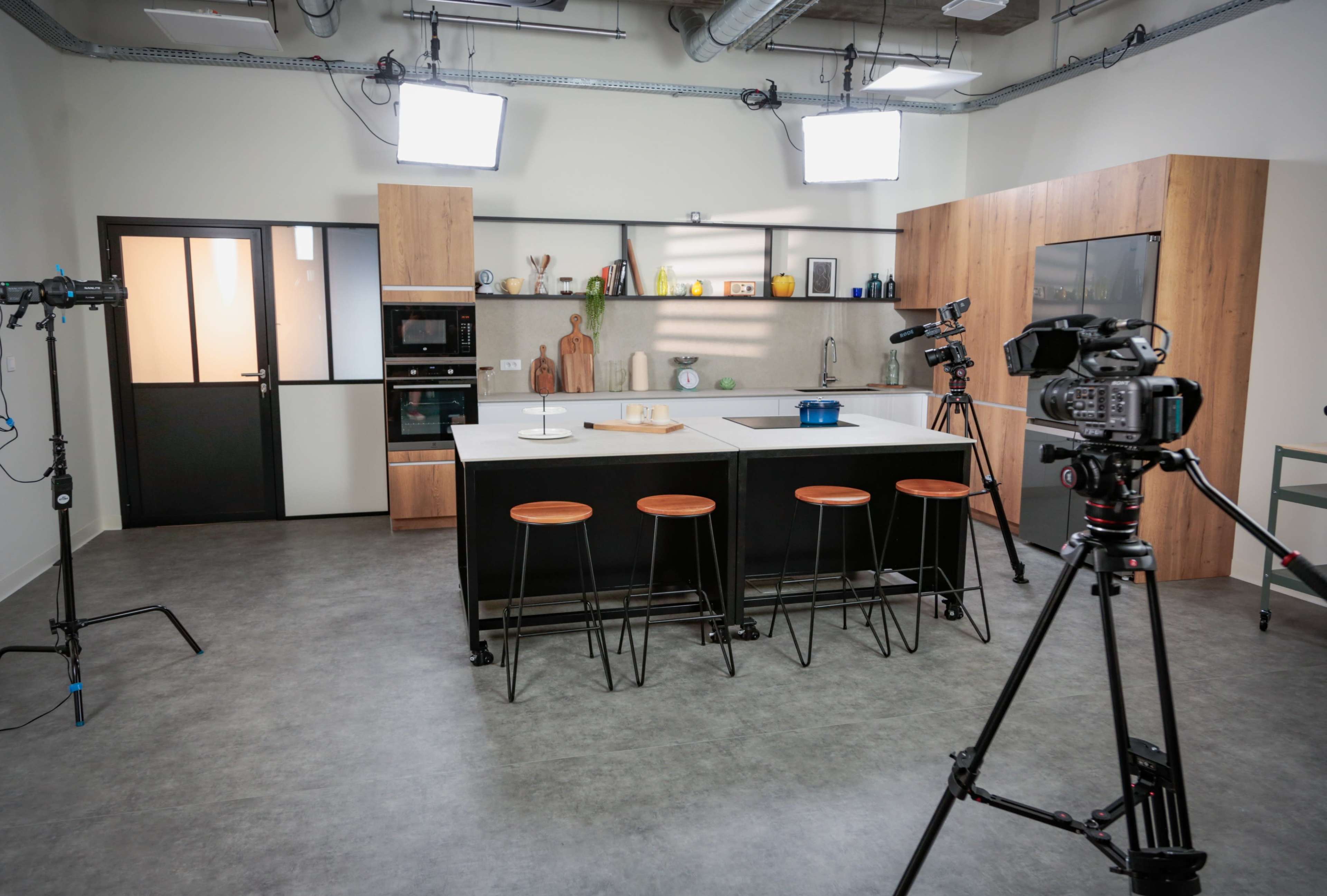 The image displays a modern kitchen studio setup with a large island, three high stools, and cameras positioned to capture the space.