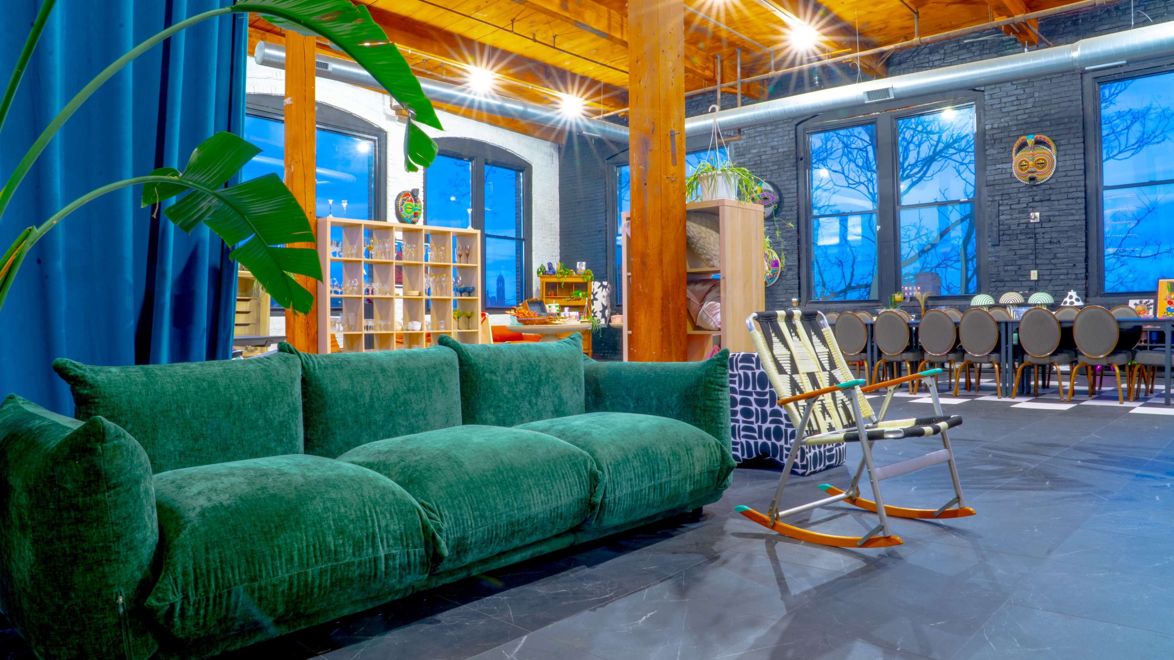 The image shows a spacious interior with a green velvet sofa, wooden beams, and a dining area in the background.