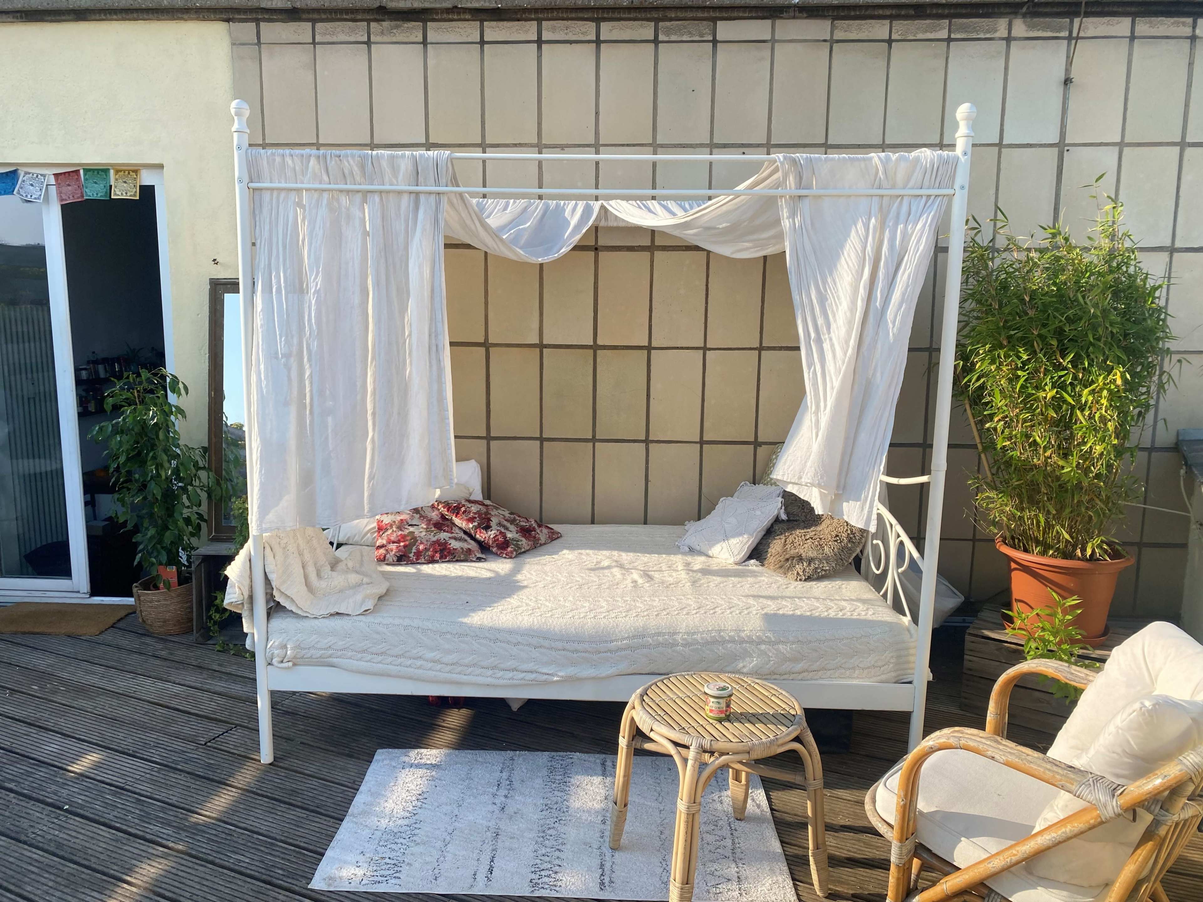 The image shows a white canopy bed set on a wooden deck, adorned with light curtains and cushions, with a potted plant nearby and a small table in front.